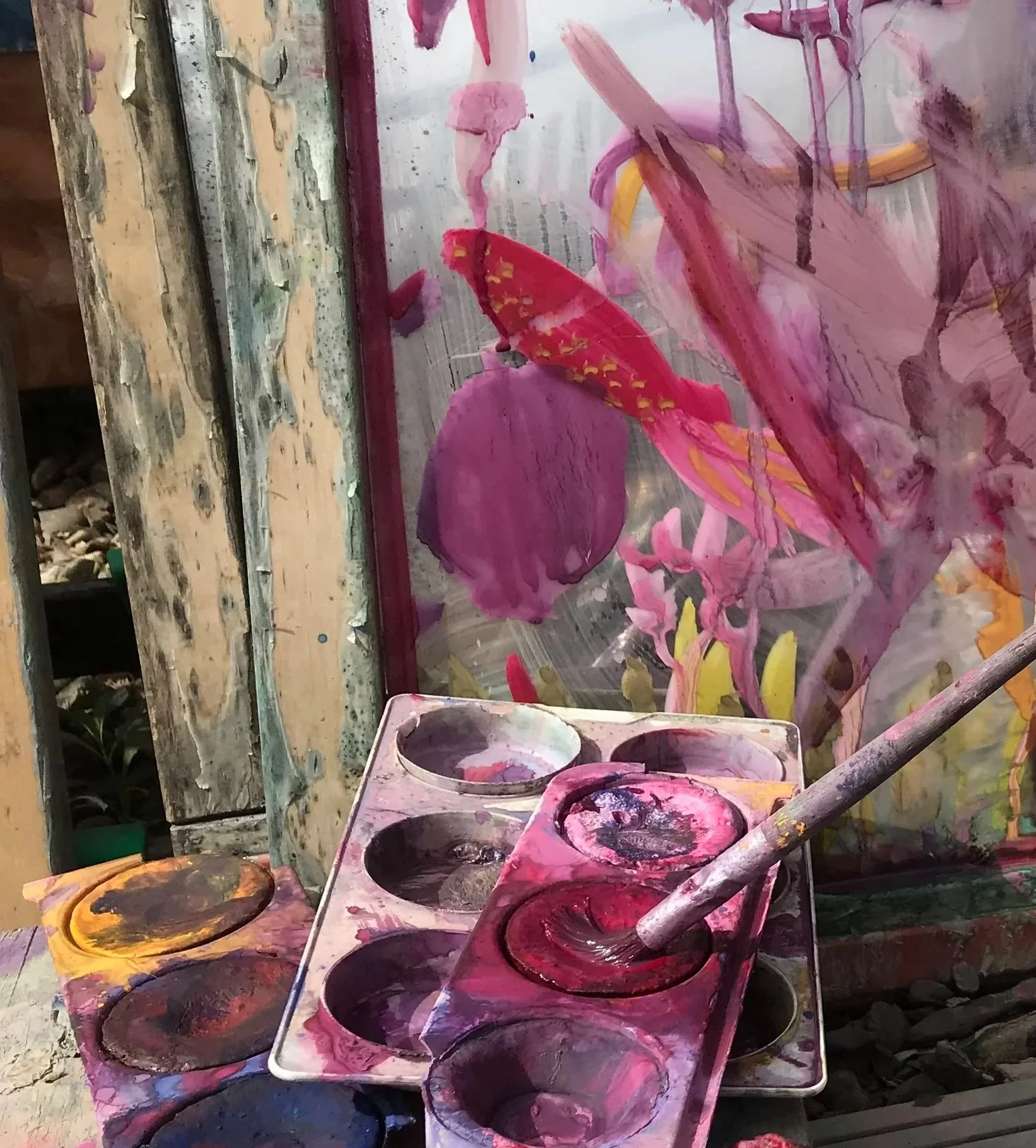 A painter's workspace with a painting in progress, featuring bold pink, purple, and yellow brushstrokes, and various paint palettes filled with dark red, black, and pink paint.