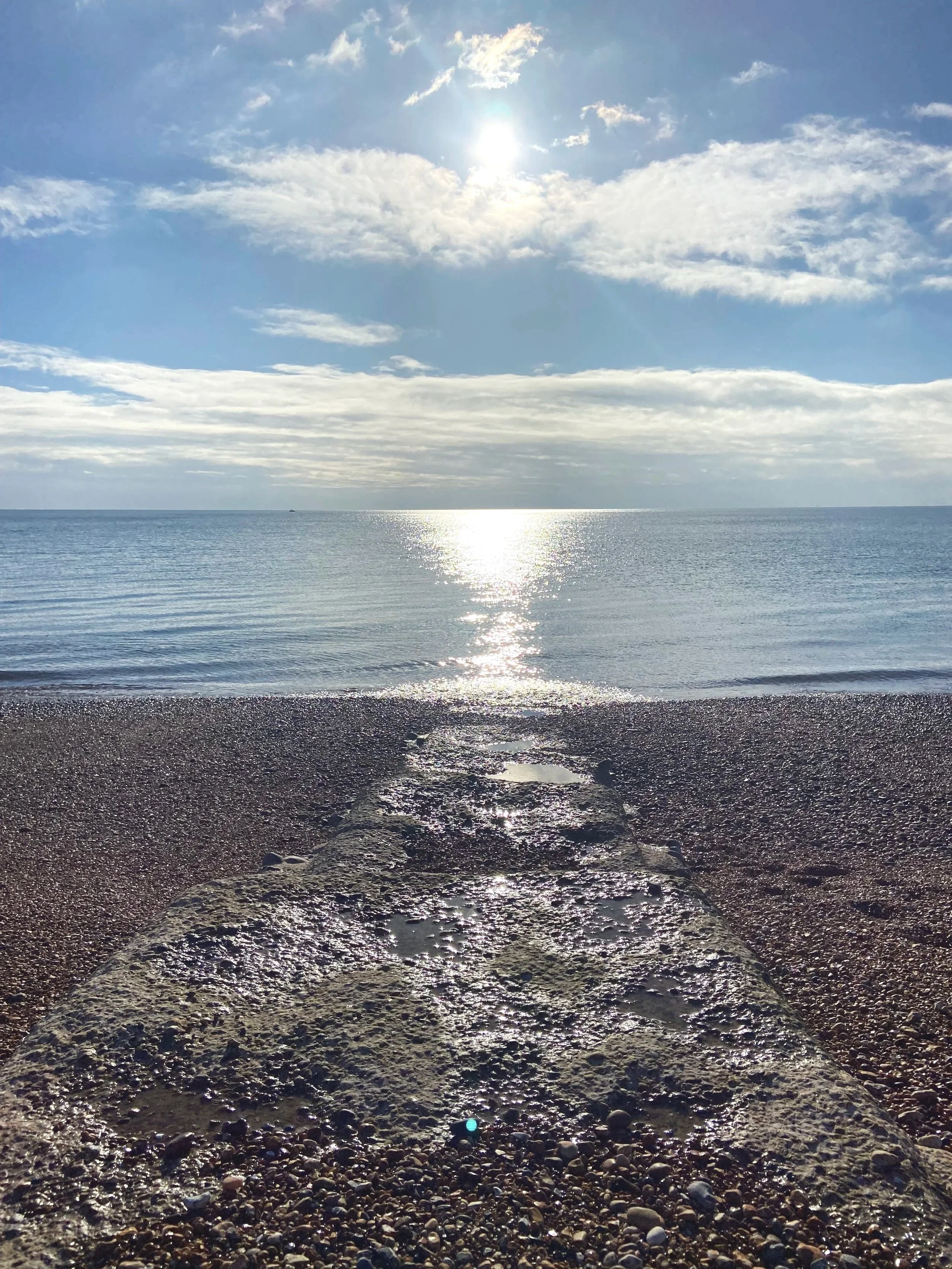 A pebble-covered beach with a concrete pier leading into the calm ocean, sun shining through partly cloudy sky overhead.