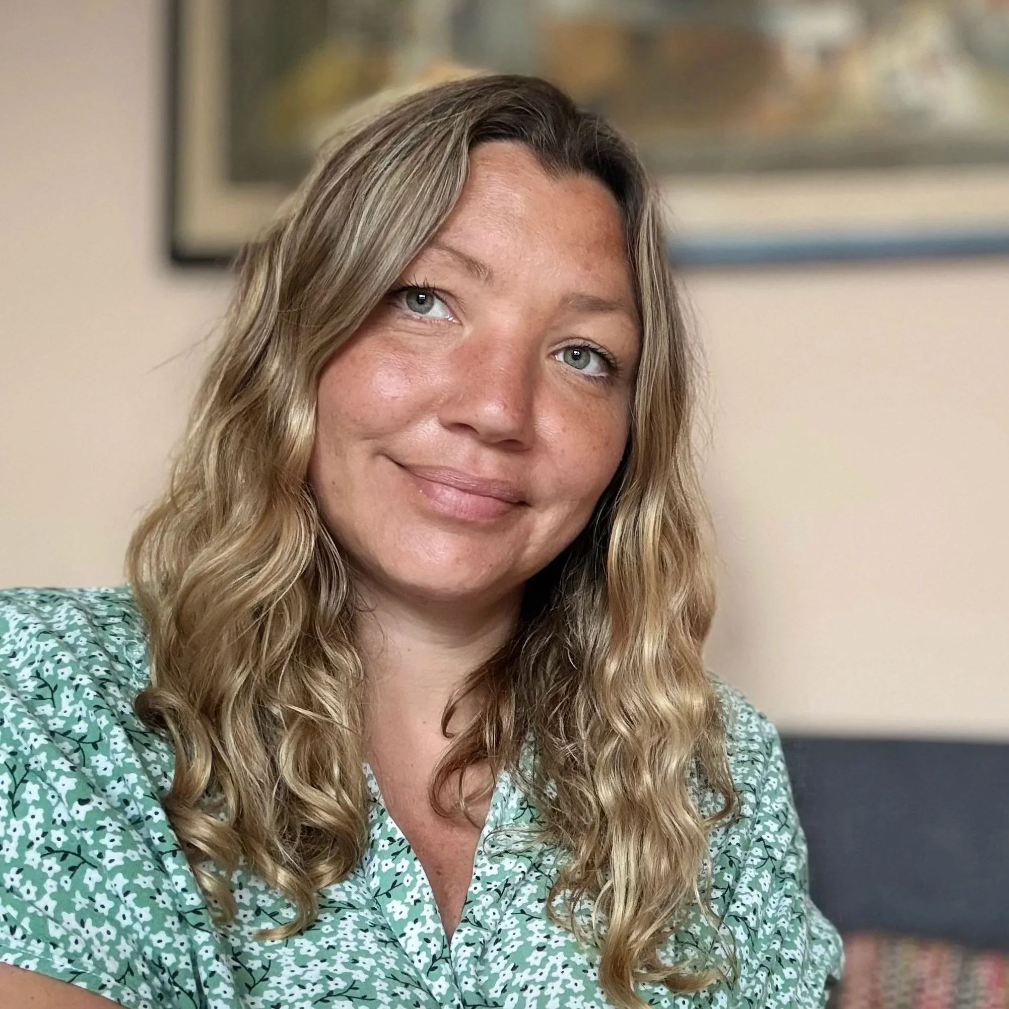 Close-up photograph of a woman with long, wavy blonde hair, smiling softly, wearing a green patterned blouse, with part of her face and upper chest visible, in an indoor setting with a blurred framed picture in the background.