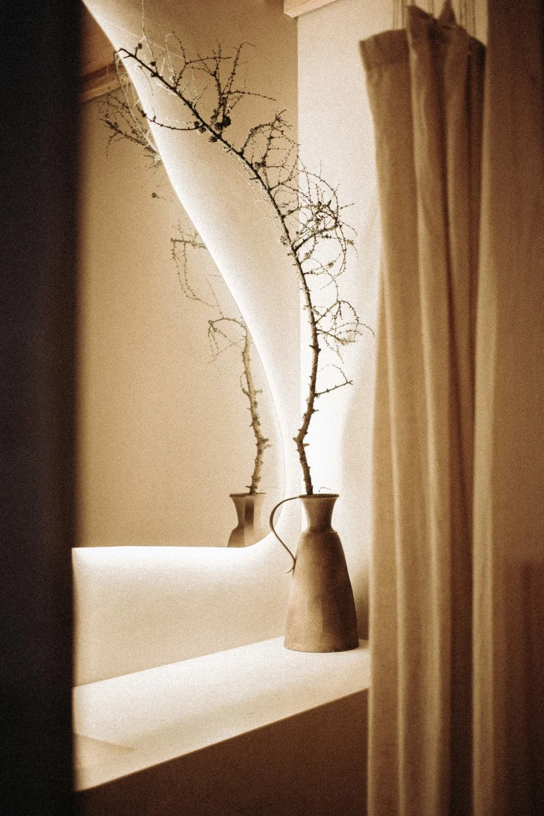 Two beige vases with bare, twisted branches are placed on a white shelf, with a mirror reflecting the scene and a curtain partially visible on the right side.