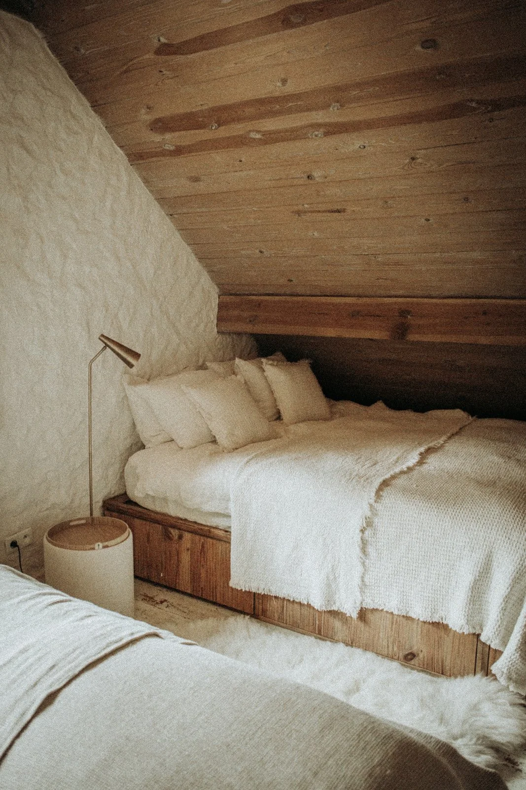 Cozy bedroom with a bed covered in white textured bedding and multiple pillows, a wooden nightstand, a modern reading lamp, and a textured wall with wooden ceiling panels.