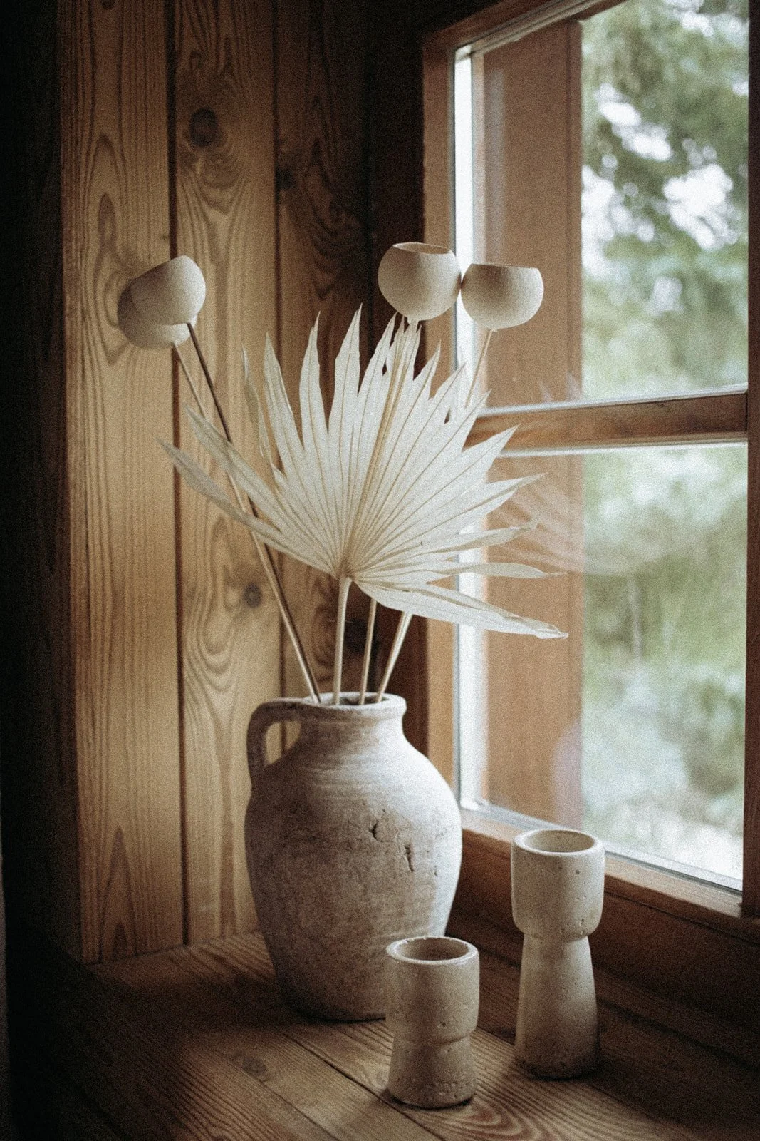 A rustic vase with dried white palm leaves and artichoke-shaped decorations on sticks on a wooden windowsill, with two small pottery cups.