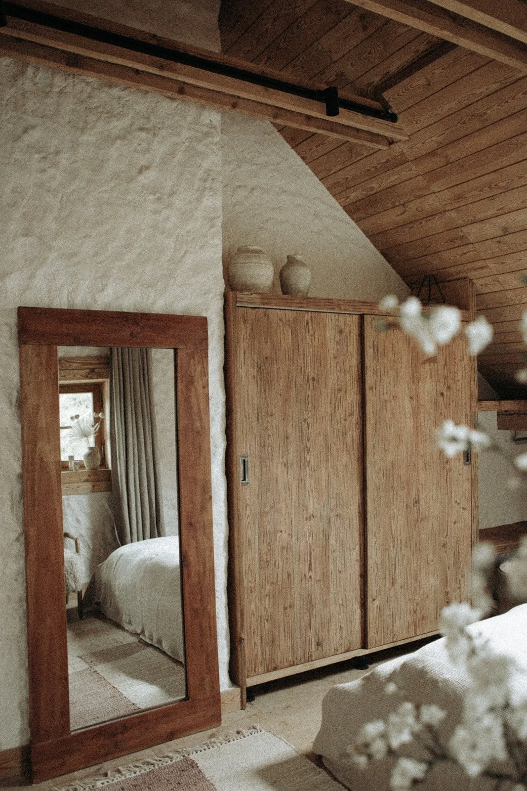 Interior of a cozy room featuring a wooden wardrobe, a full-length mirror with a wooden frame, a bed with white linens, a window with beige curtains, and decorative pottery on top of the wardrobe, with wooden paneling on the ceiling.
