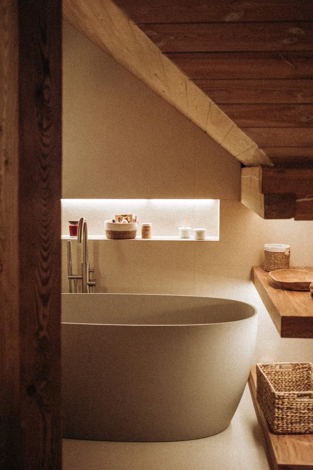 A modern bathroom featuring a freestanding bathtub under a sloped wooden ceiling. The space includes a shelf with containers and decor items, a wooden countertop with a wicker basket, and woven storage cubbies.