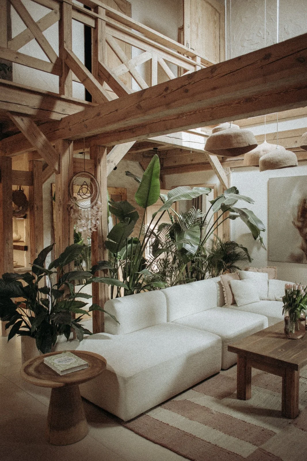 Living room with white sectional sofa, wooden coffee table with a vase of pink tulips, surrounded by large green plants, wooden beams, and modern hanging lamps.