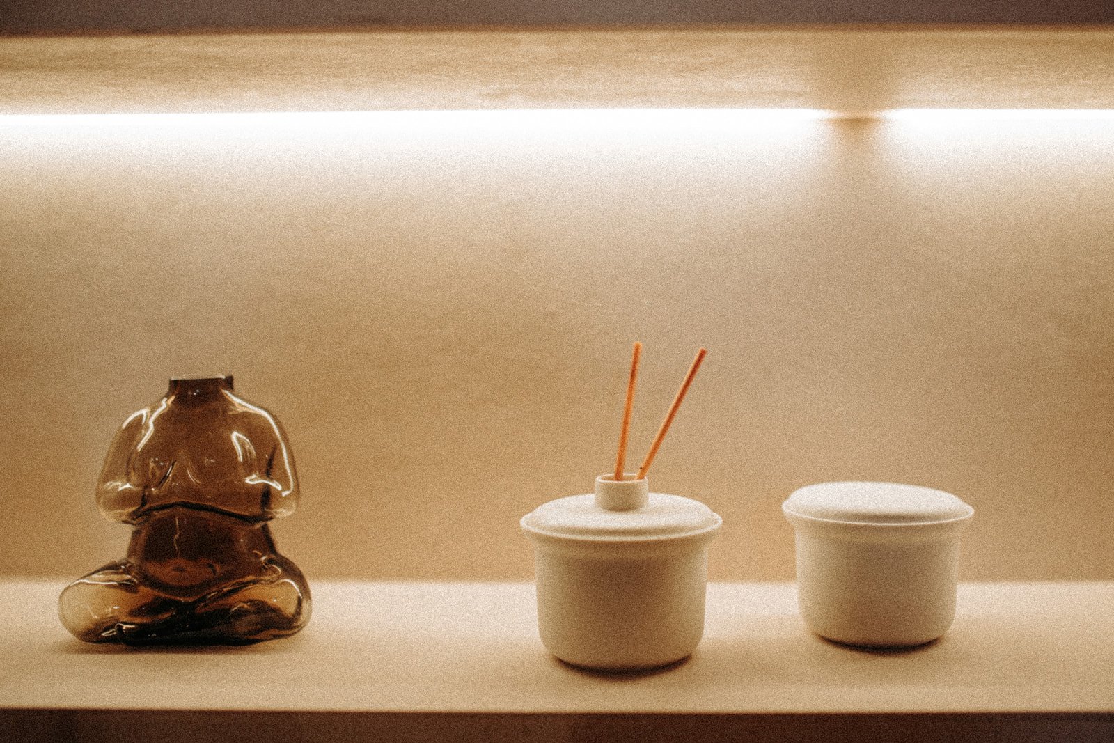 A small display shelf with three decorative items: a dark brown, glossy ceramic figurine of a meditative monk, a white ceramic container with two orange chopsticks sticking out, and a closed white ceramic container.