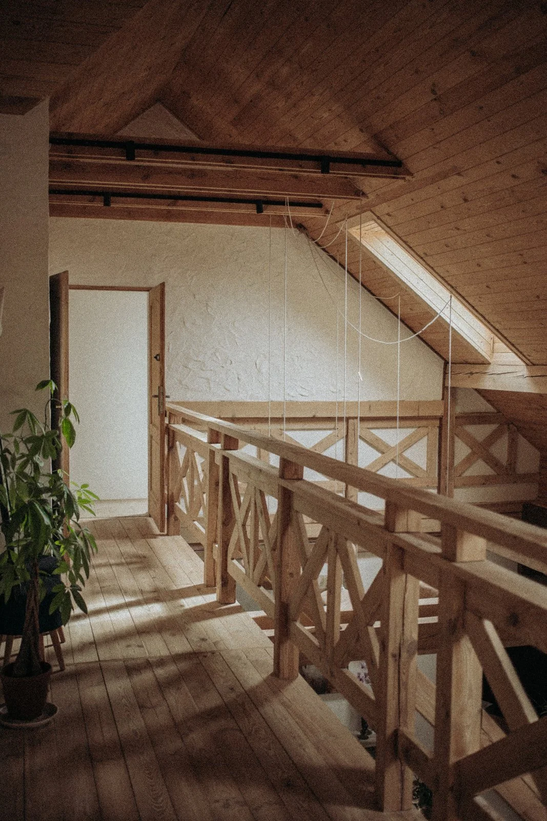 Interior view of a wooden loft space with a wooden staircase and railing, a white textured wall, an open door, a plant on the floor, and a sloped wooden ceiling.