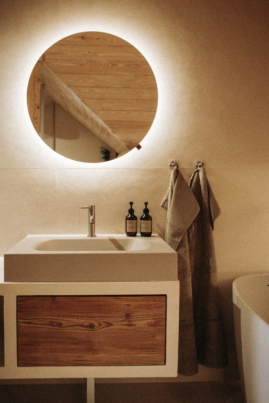Bathroom with a round mirror, wooden cabinet under the sink, two hand towels hanging on hooks, and two pump bottles on the sink. Warm lighting.