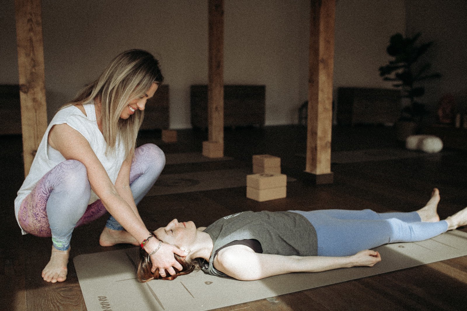 A woman practicing yoga or giving a relaxation massage to another woman lying on the mat in a dimly lit, cozy indoor space with wooden beams and minimal decor.