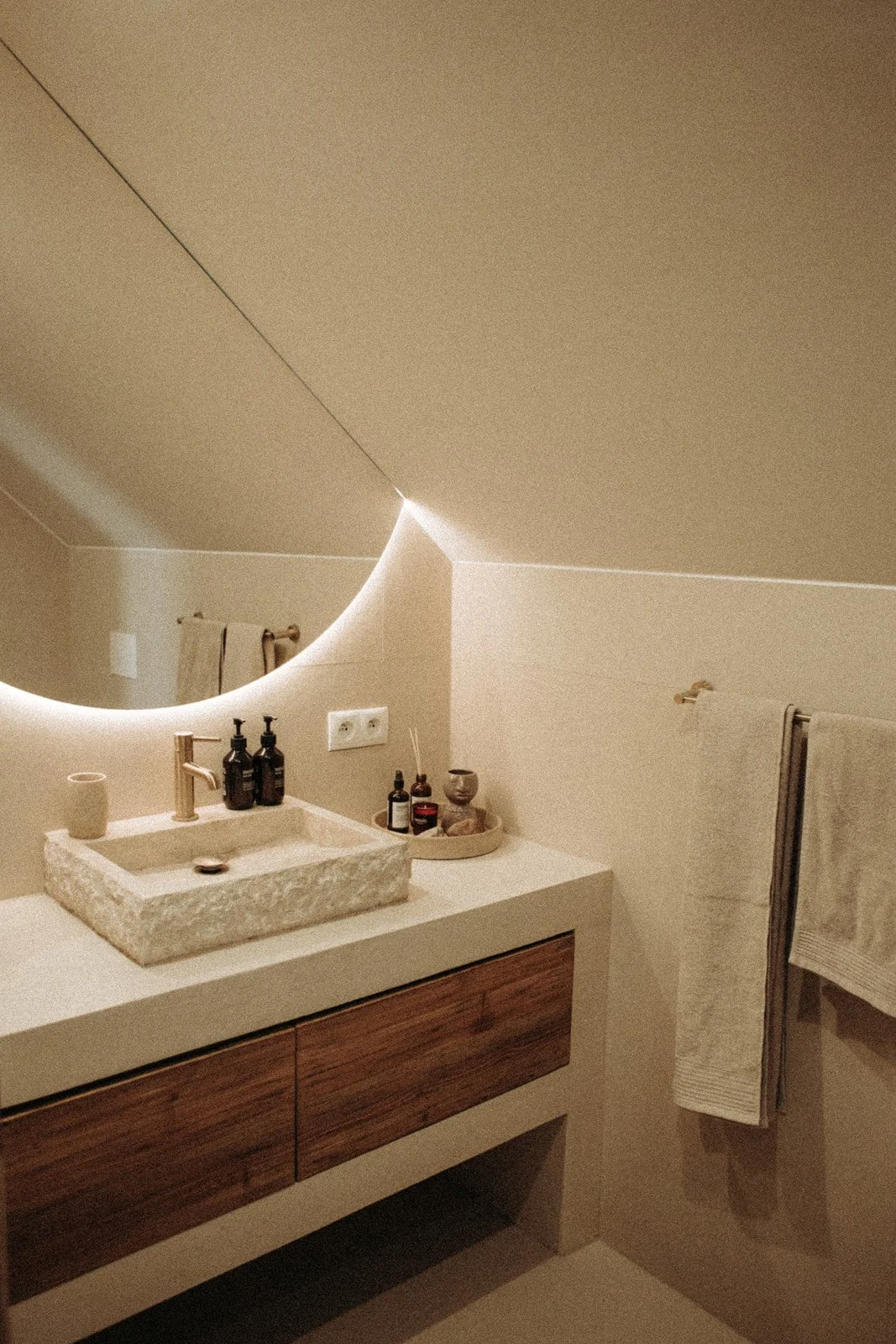 Modern bathroom vanity with stone sink, black soap dispensers, small decorative items, and a towel hanging on a rack next to a circular mirror with backlighting.