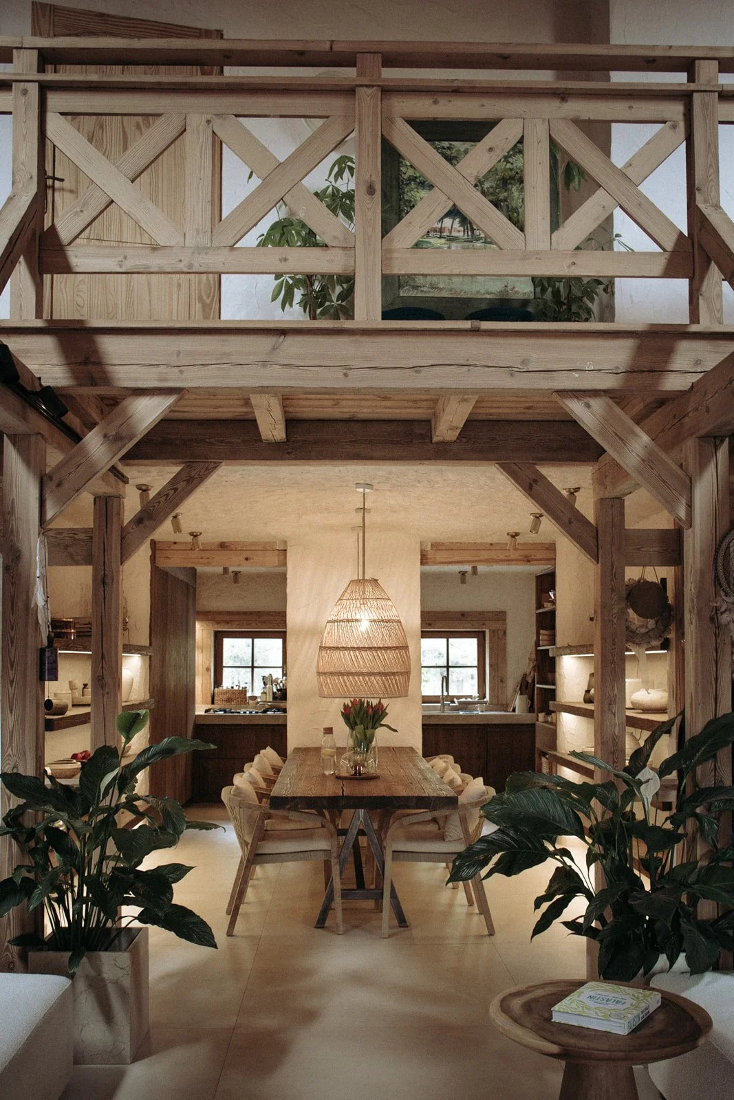 A dining area featuring a rustic wooden table with eight chairs, a hanging pendant light, a vase with tulips, and large potted plants, with a view of a kitchen in the background.