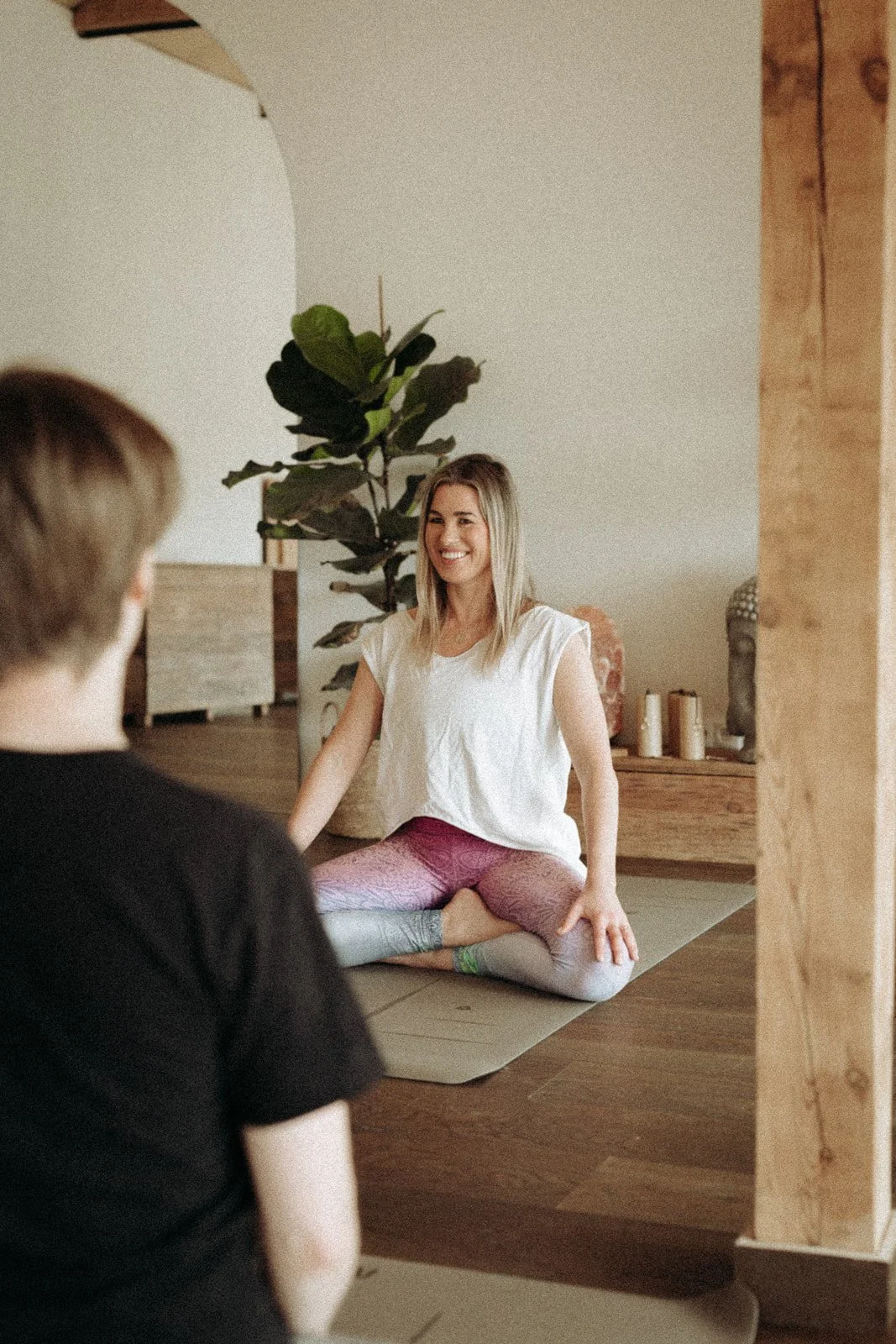 A woman practicing yoga or meditation indoors, sitting cross-legged on a yoga mat, smiling, with a potted plant and candles in the background.
