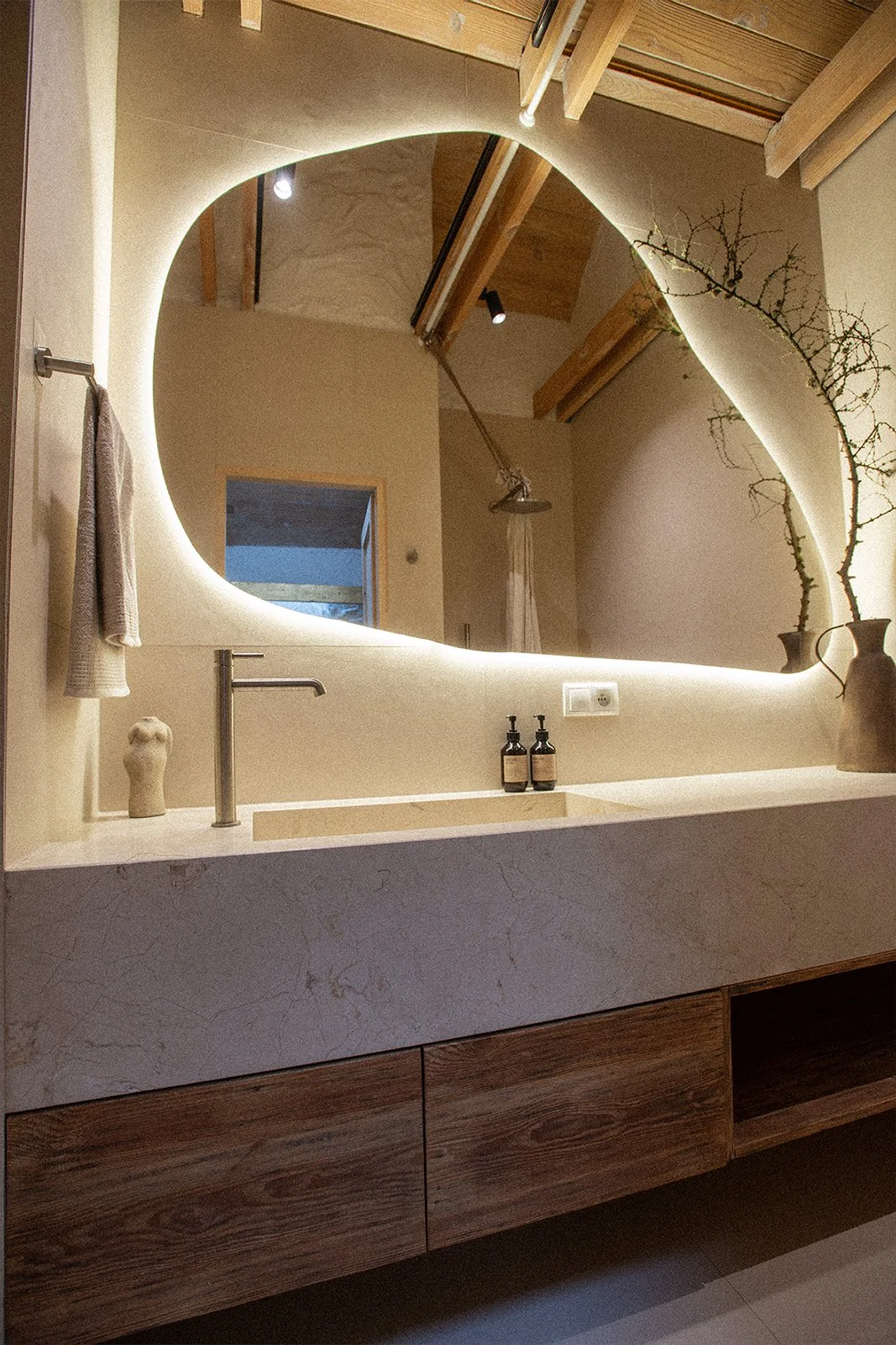 A modern bathroom vanity with a large, backlit, irregularly shaped mirror, a beige countertop with a built-in sink, two soap dispensers, and decorative vases with branches. A towel hangs on a hook beside the mirror. The background shows a wooden ceil