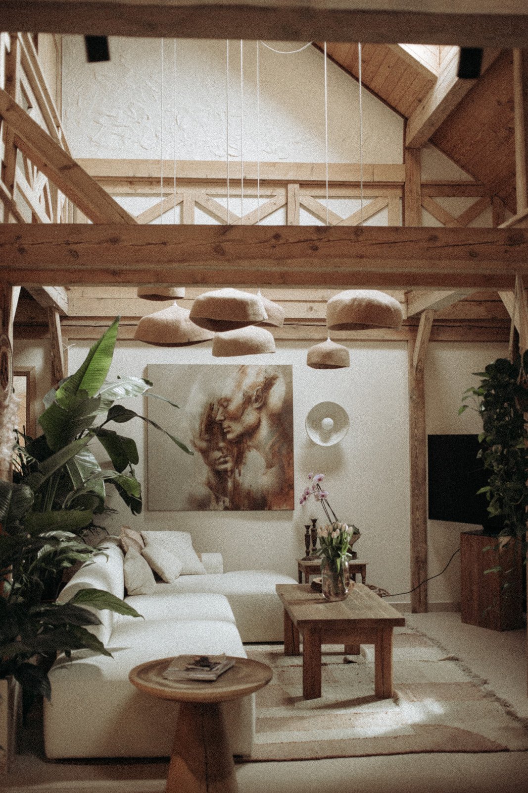 Living room with white sofa, wooden coffee table, large green plants, abstract art on the wall, and hanging clay lamps under a wooden ceiling with exposed beams.