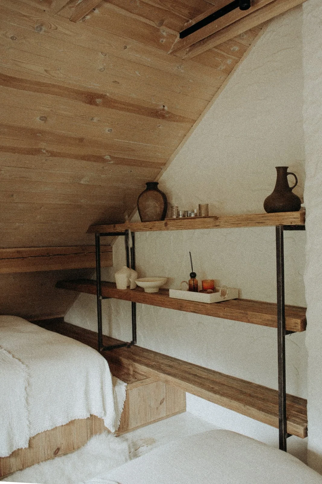 A corner of a bedroom with wooden ceiling and shelves, a bed with white blanket, and decorative vases and objects on the shelves.