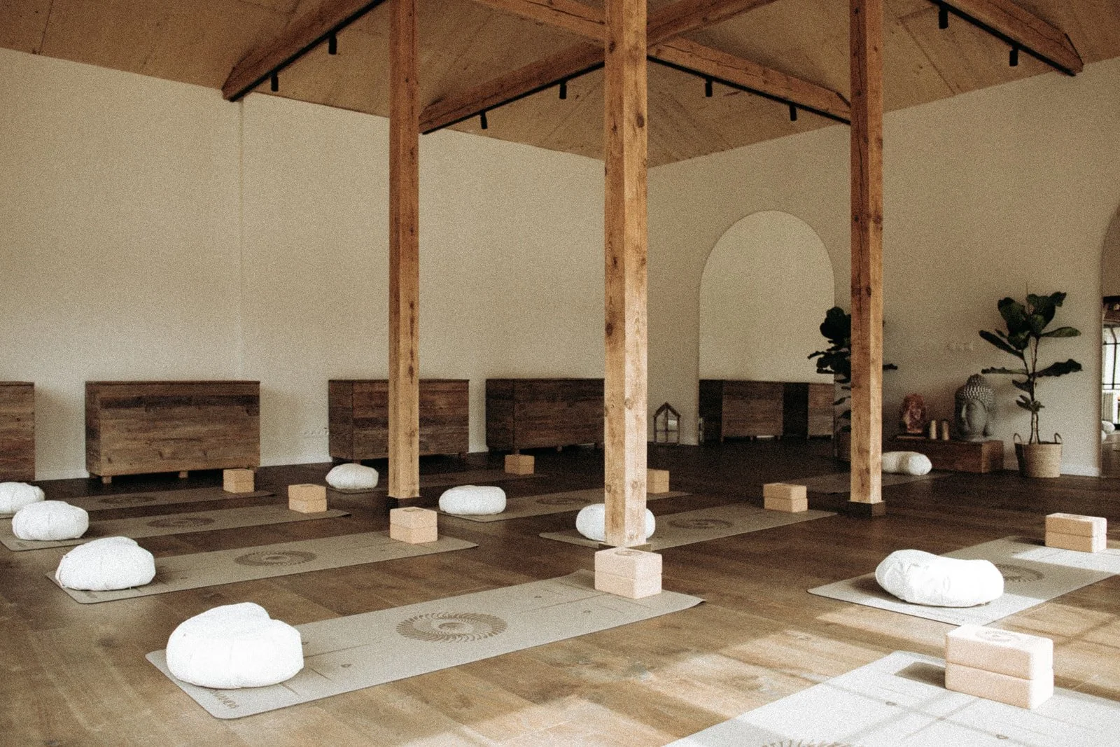 A serene yoga studio with wooden floors and beams, equipped with mats, bolsters, and blocks for meditation or yoga practice. Potted plants and decorative items decorate the space, creating a calm, natural atmosphere.
