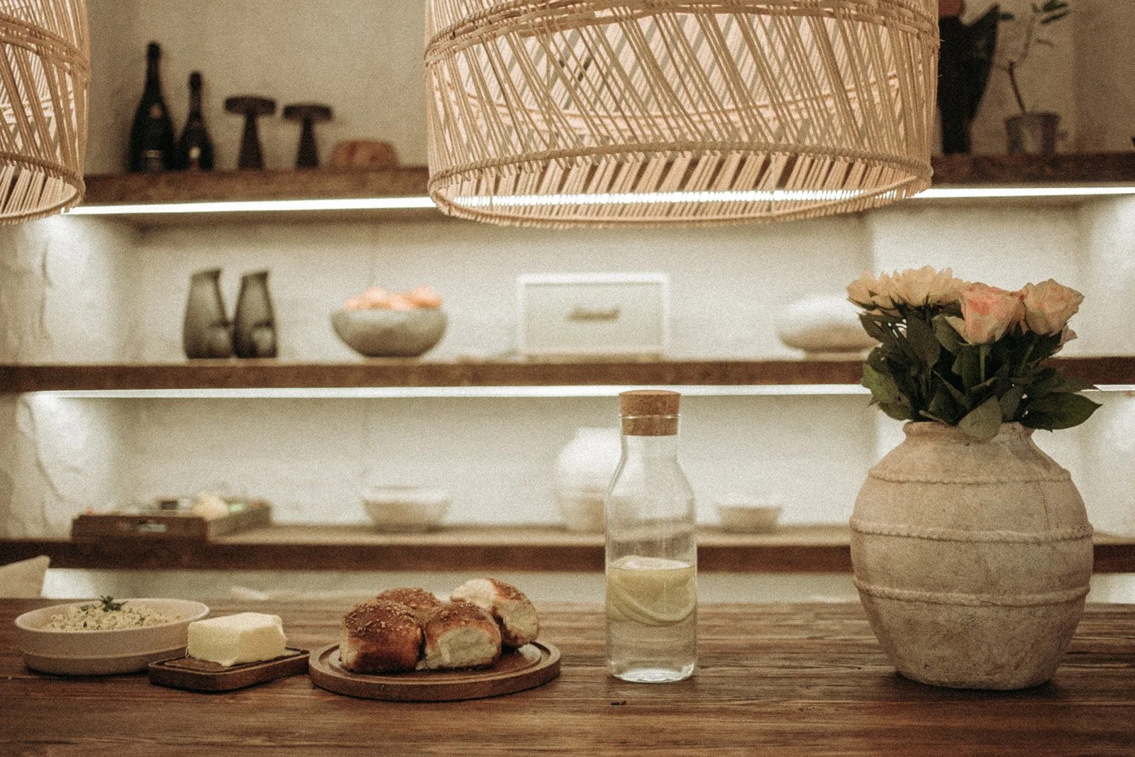 A wooden dining table with a small bowl of salad, a pat of butter, a plate of bread rolls, a glass bottle of water, and a vase with pink roses. In the background, shelves with decorative bottles, bowls, and other items, illuminated by warm light and 