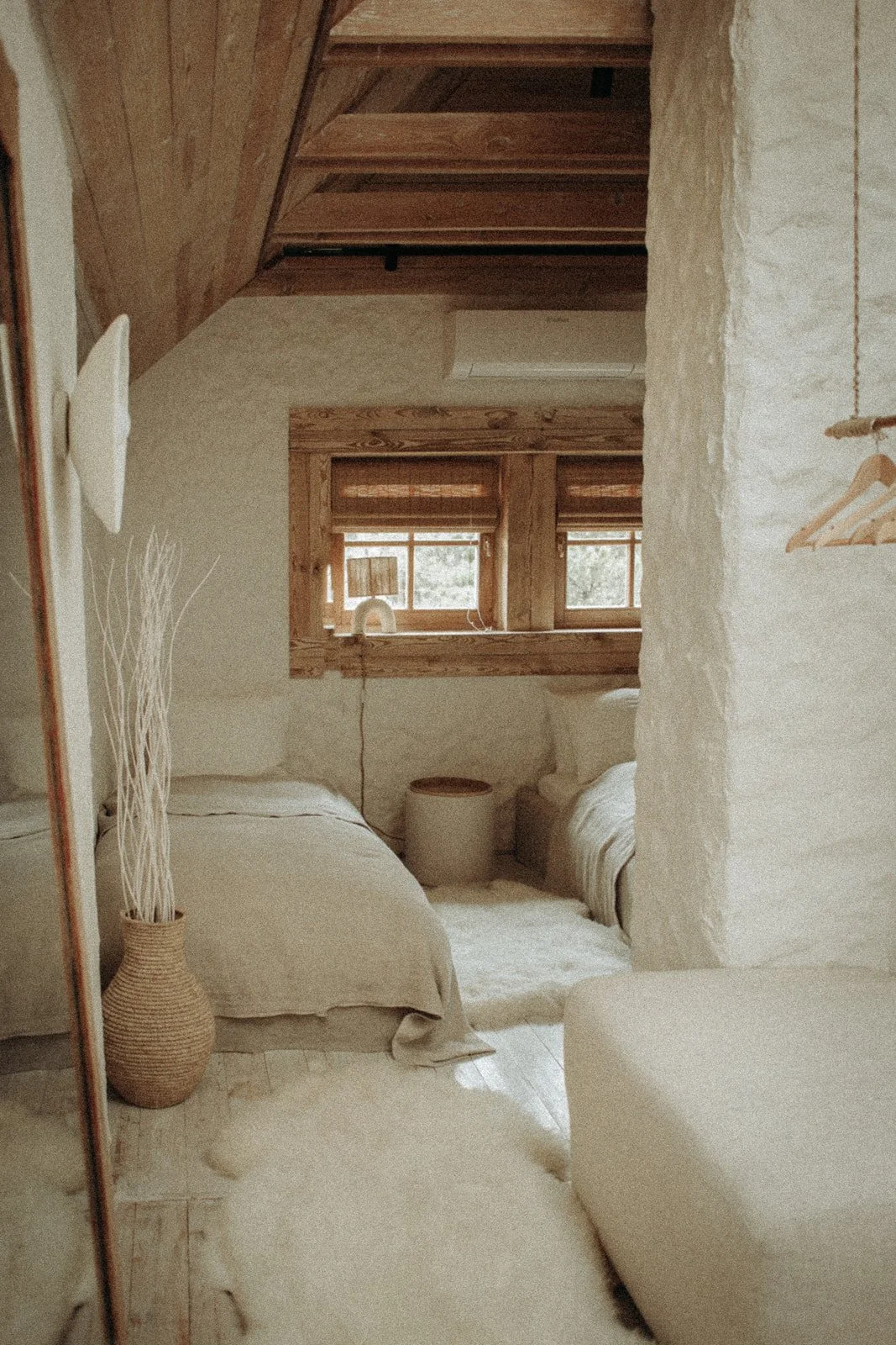 A cozy bedroom with beige bedding and wooden accents. Two small windows with blinds, a round side table, and minimalist decor including a tall wicker vase with dried plants.