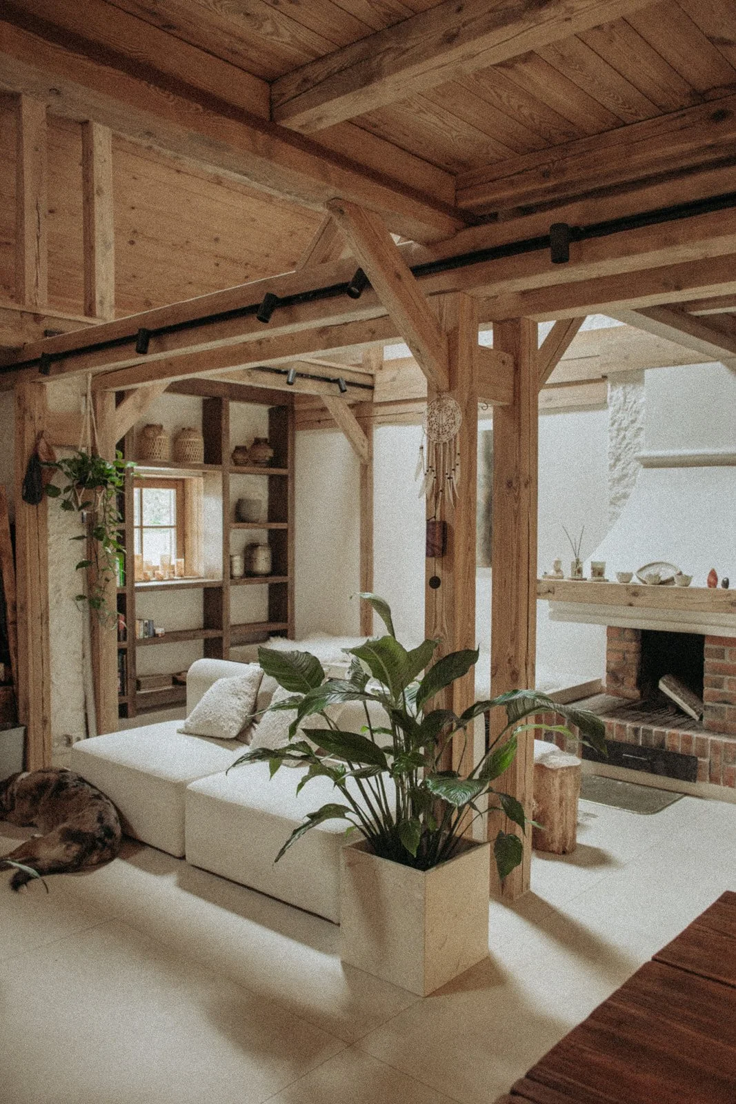 Salon w Ananda Retreat House z drewnianymi belkami, jasną sofą, kominkiem i naturalnymi elementami wystroju.