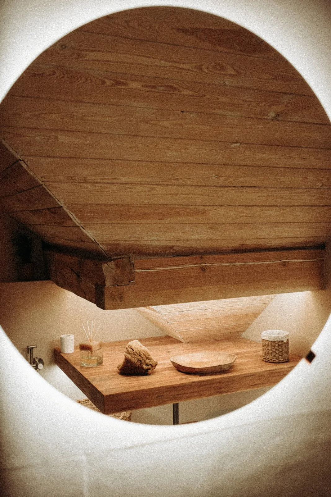 A wooden shelf with decorative objects, including a rock, a wooden bowl, a reed diffuser, a candle, and a wicker basket, seen through a circular opening in a wall.