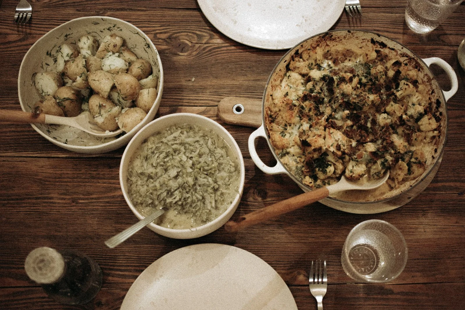 A table set with a casserole dish of baked dish, a bowl of cooked potatoes with herbs, a bowl of chopped cabbage, and some empty plates and glasses.