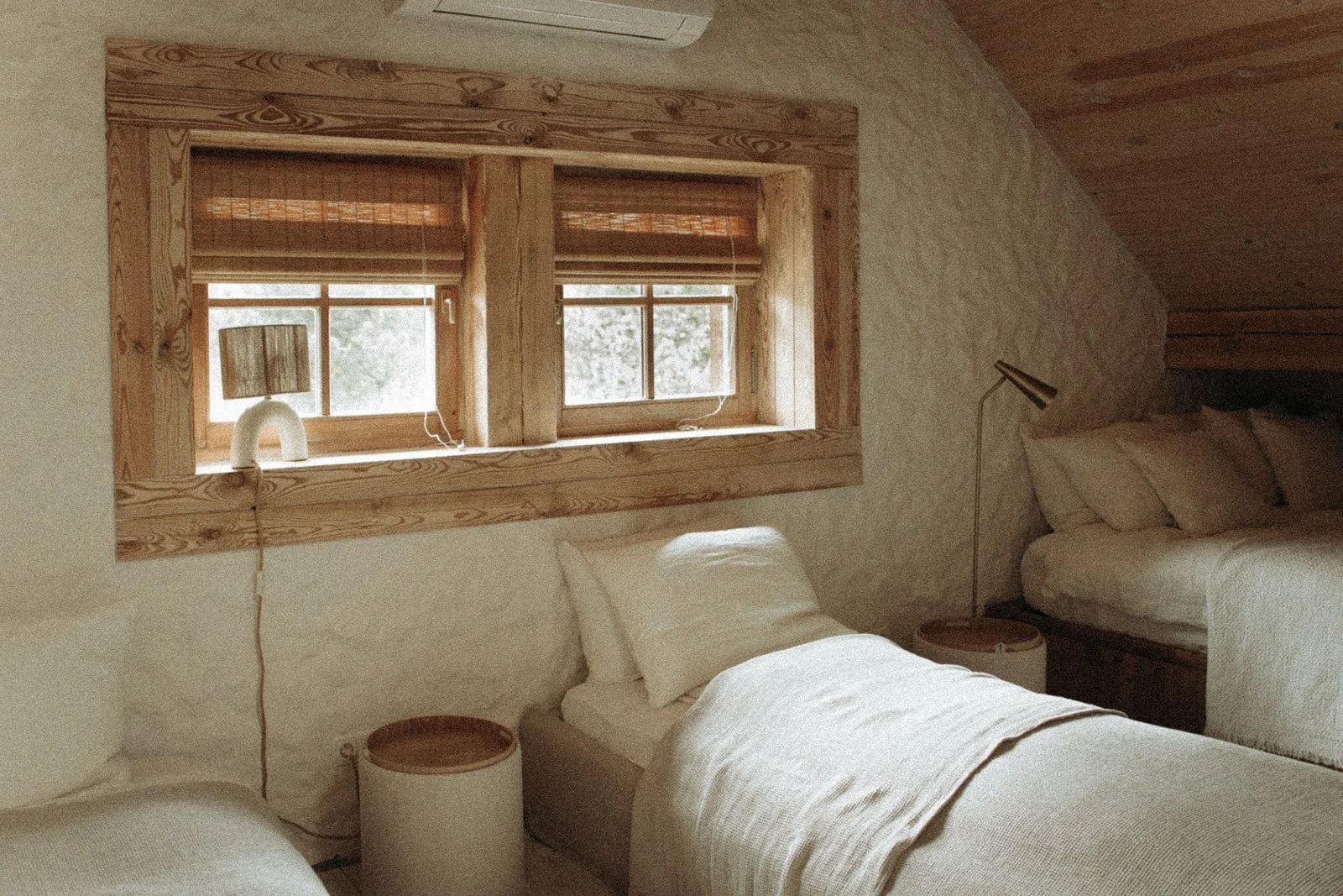 A cozy bedroom with two beds, a window with wooden framing, and a lamp on a side table next to the bed. The room has a rustic, natural decor style.