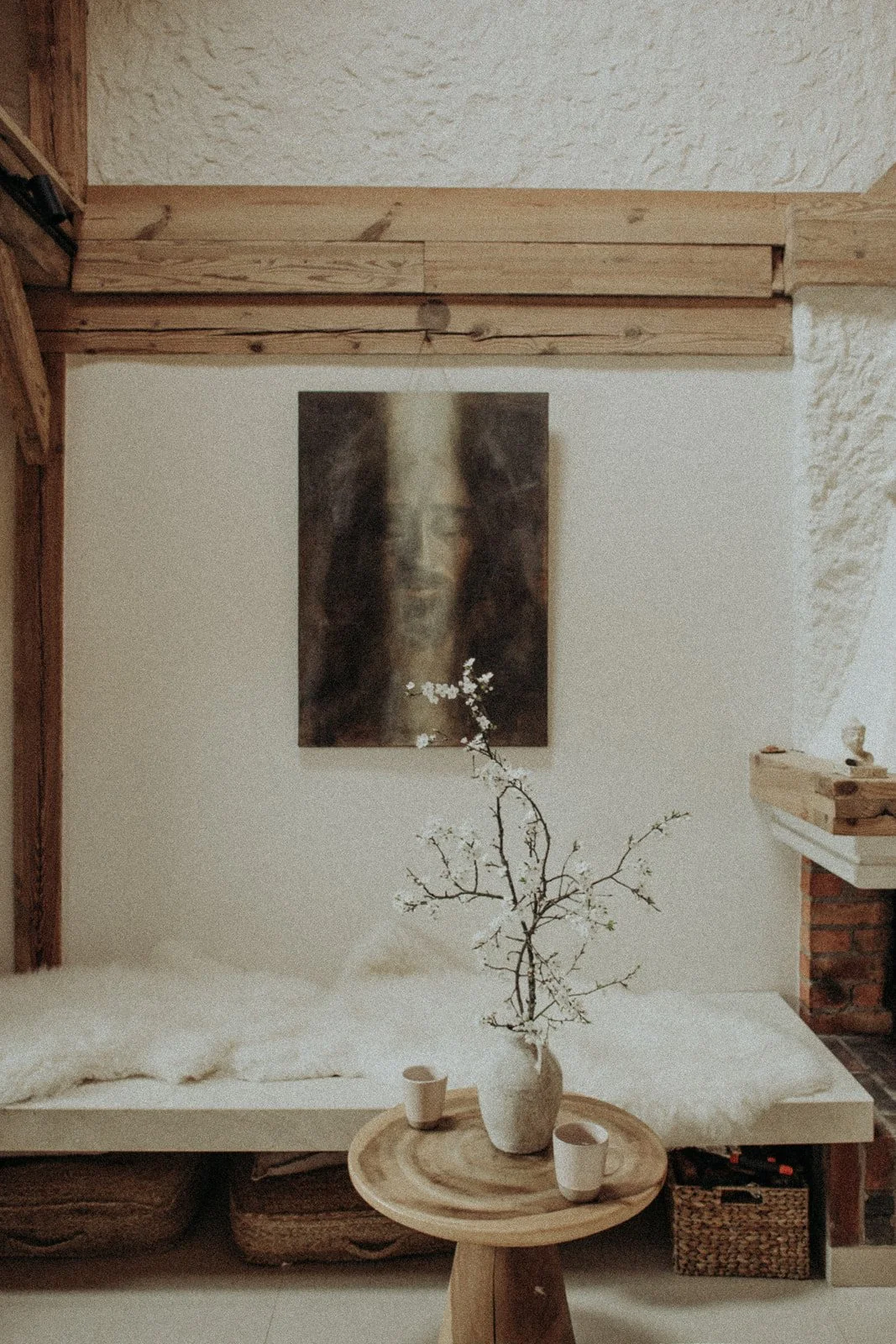A cozy living room corner featuring a white couch with a throw blanket, a round wooden table with a white vase holding a branch with white blossoms, and two white cups. Behind the table, a plush blanket is draped, and a woven basket is visible undern