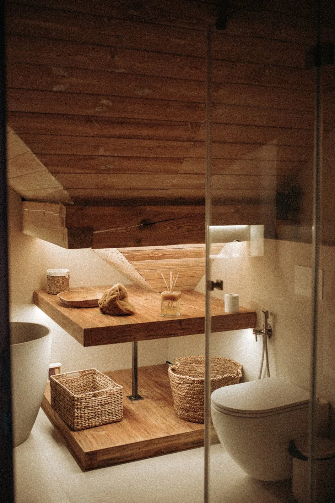A cozy bathroom with a wooden sloped ceiling, a wooden shelf, and baskets. The shelf has a small bowl, a fur item, and a reed diffuser. There is a toilet with a toilet paper roll and a handheld bidet spray. The bathroom is lit softly, creating a warm