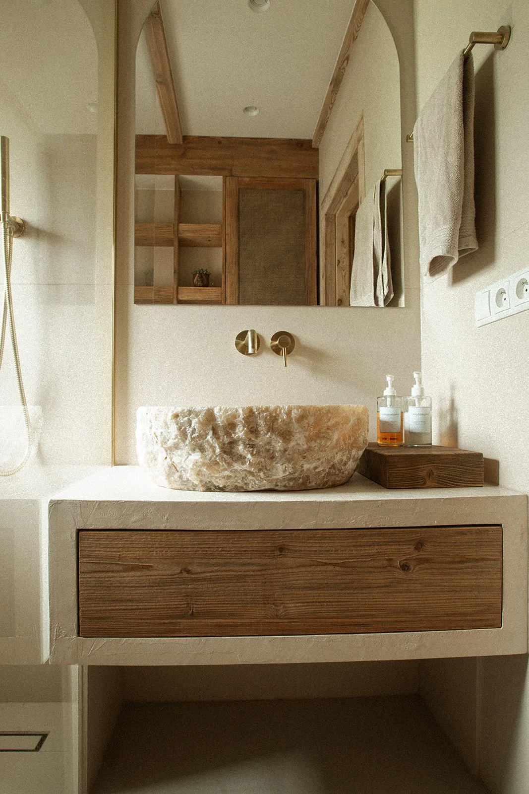 A bathroom vanity with a round stone sink, a large mirror above, and a wooden drawer beneath. There are soap dispensers on a small wooden tray to the right of the sink, and a towel hanging on a gold towel rack. The background shows a wooden cabinet a