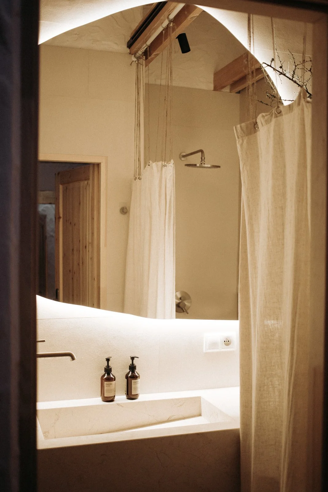 The photo shows a minimalist bathroom with a large mirror, two brown bottles of soap or lotion on a white sink, beige curtains, a shower head, and a small sink installed above the main sink. The bathroom features warm lighting and wooden accents, inc