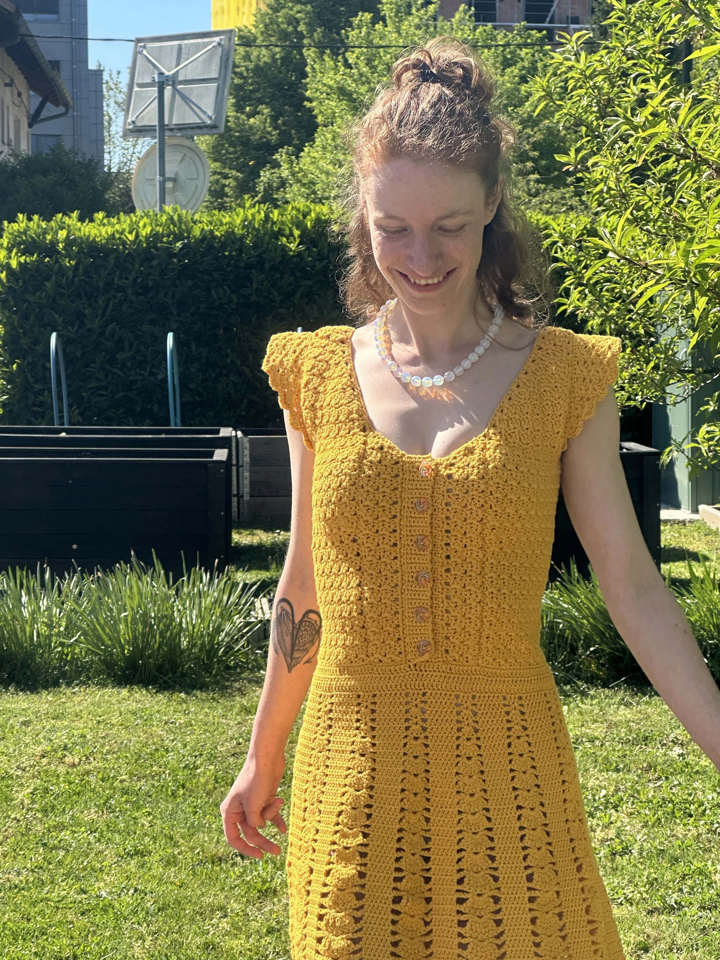 A woman with curly hair tied up in a bun, wearing a yellow crochet dress with buttons down the front, smiling outdoors on a sunny day, surrounded by greenery and trees.