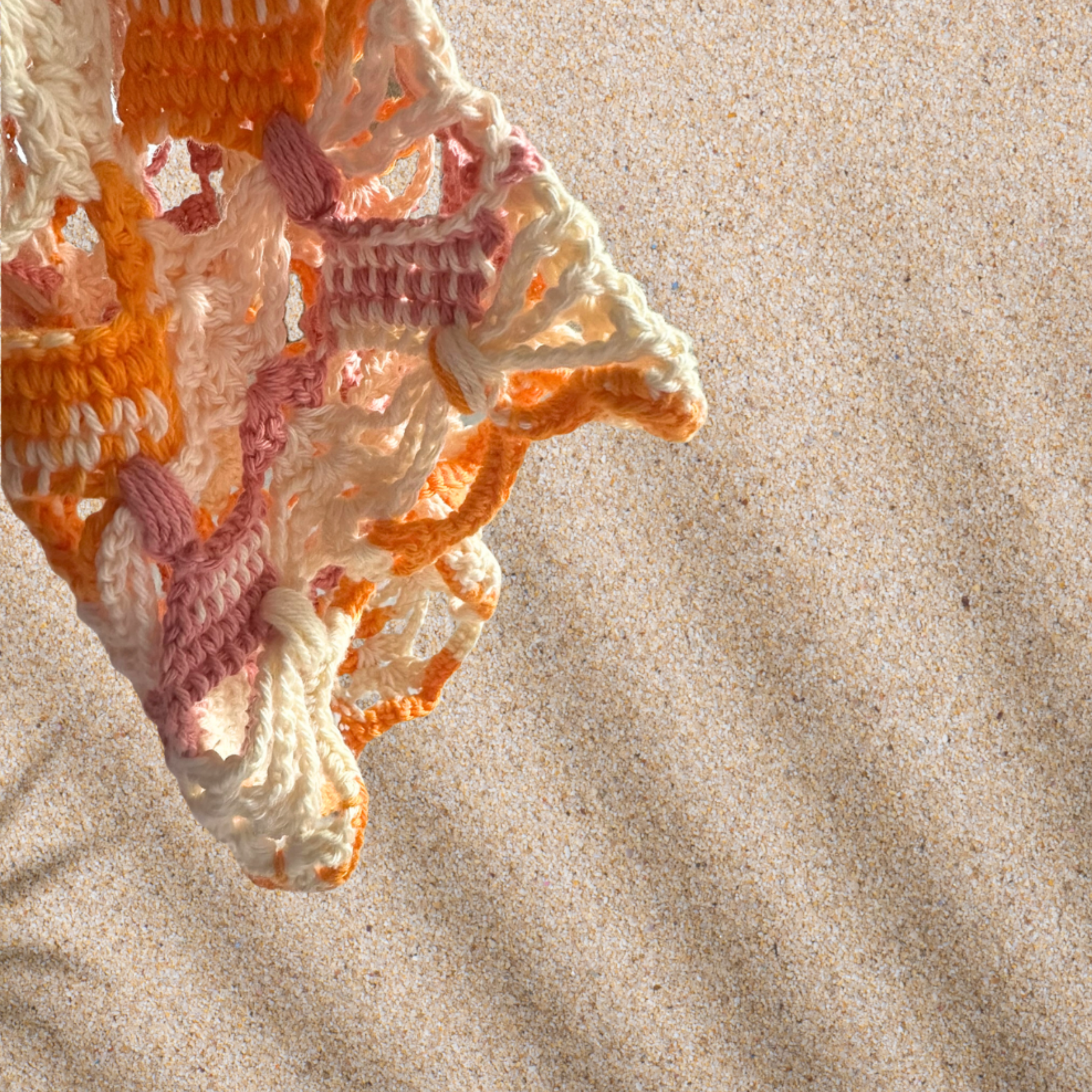 Close-up view of a colorful crocheted item with yarn in shades of white, pink, orange, and peach, lying on beige sand.