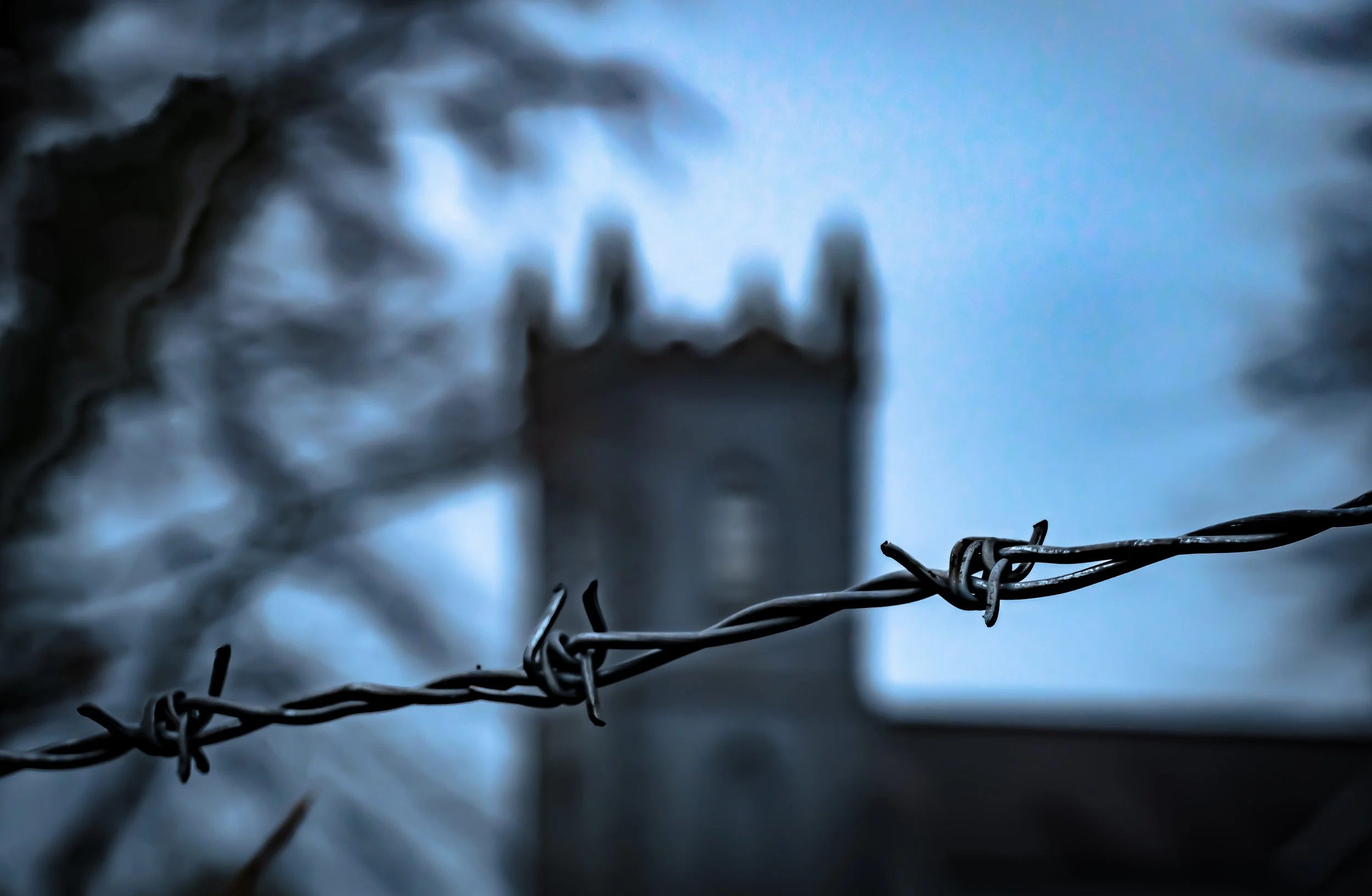 Close-up of barbed wire fence with a blurry background of a church and cloudy sky