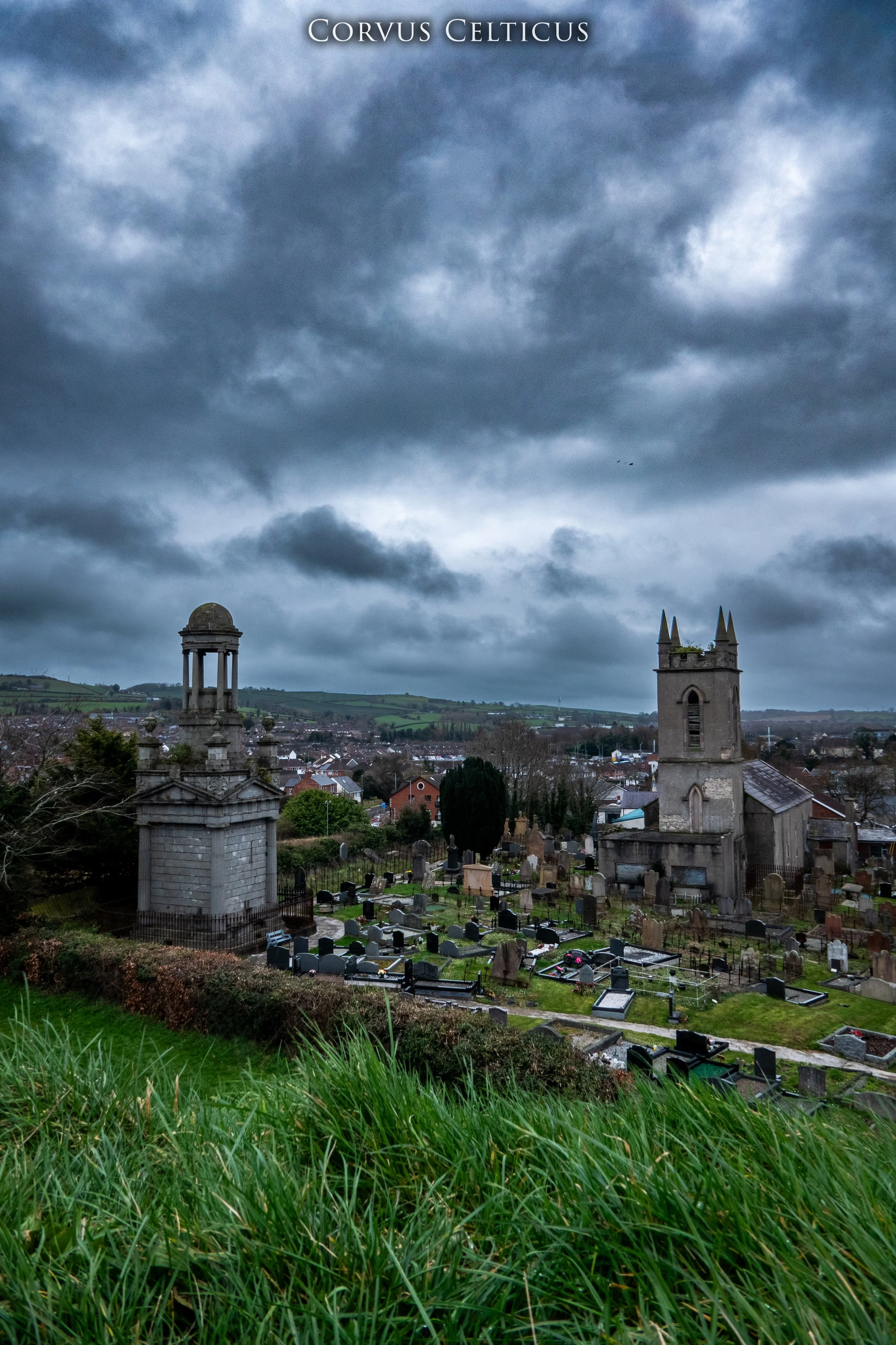 A dramatic, cloudy sky over a cemetery with two prominent towers, one being the Cleland Memorial Mausoleum and the other Saint Elizabeth's Old Church in Dundonald