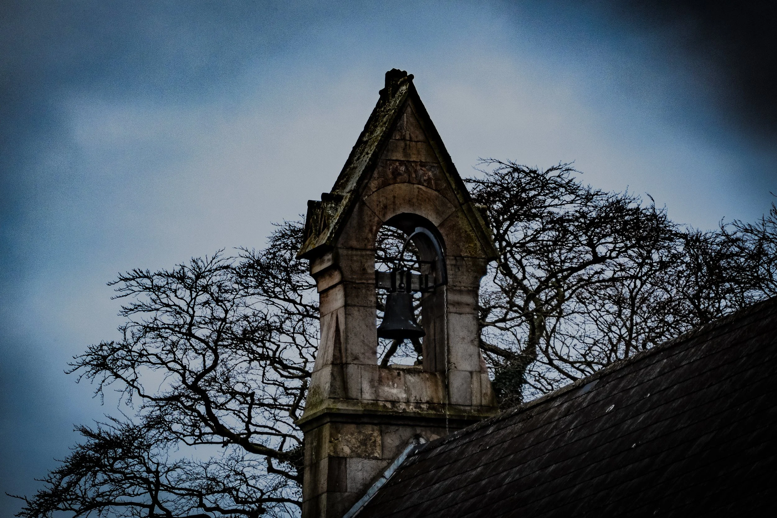 An old stone church bell tower with a bell hanging inside, surrounded by leafless trees under a cloudy sky.