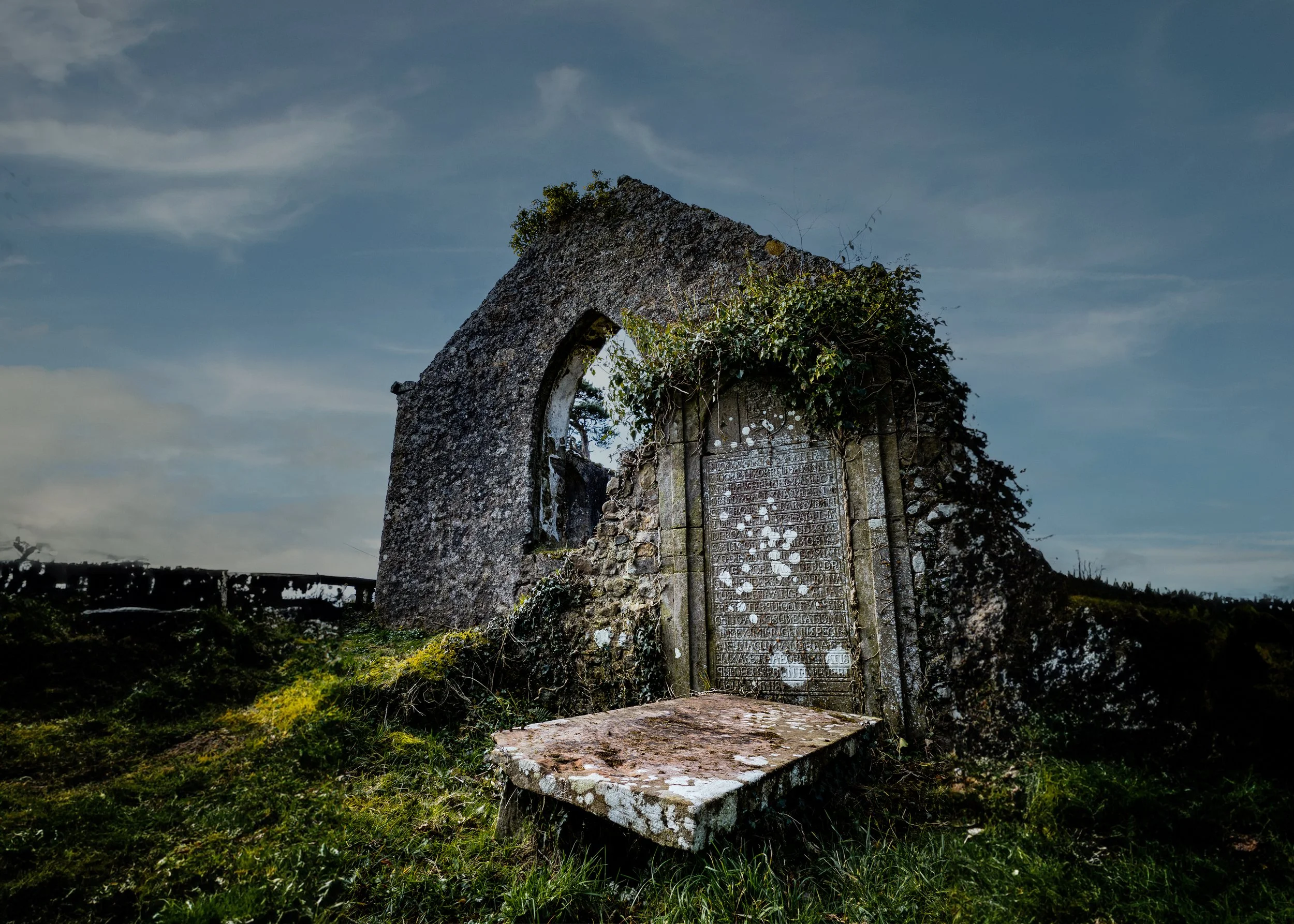 Old stone monument with a plaque containing Latin, overgrown with ivy, under a cloudy sky.