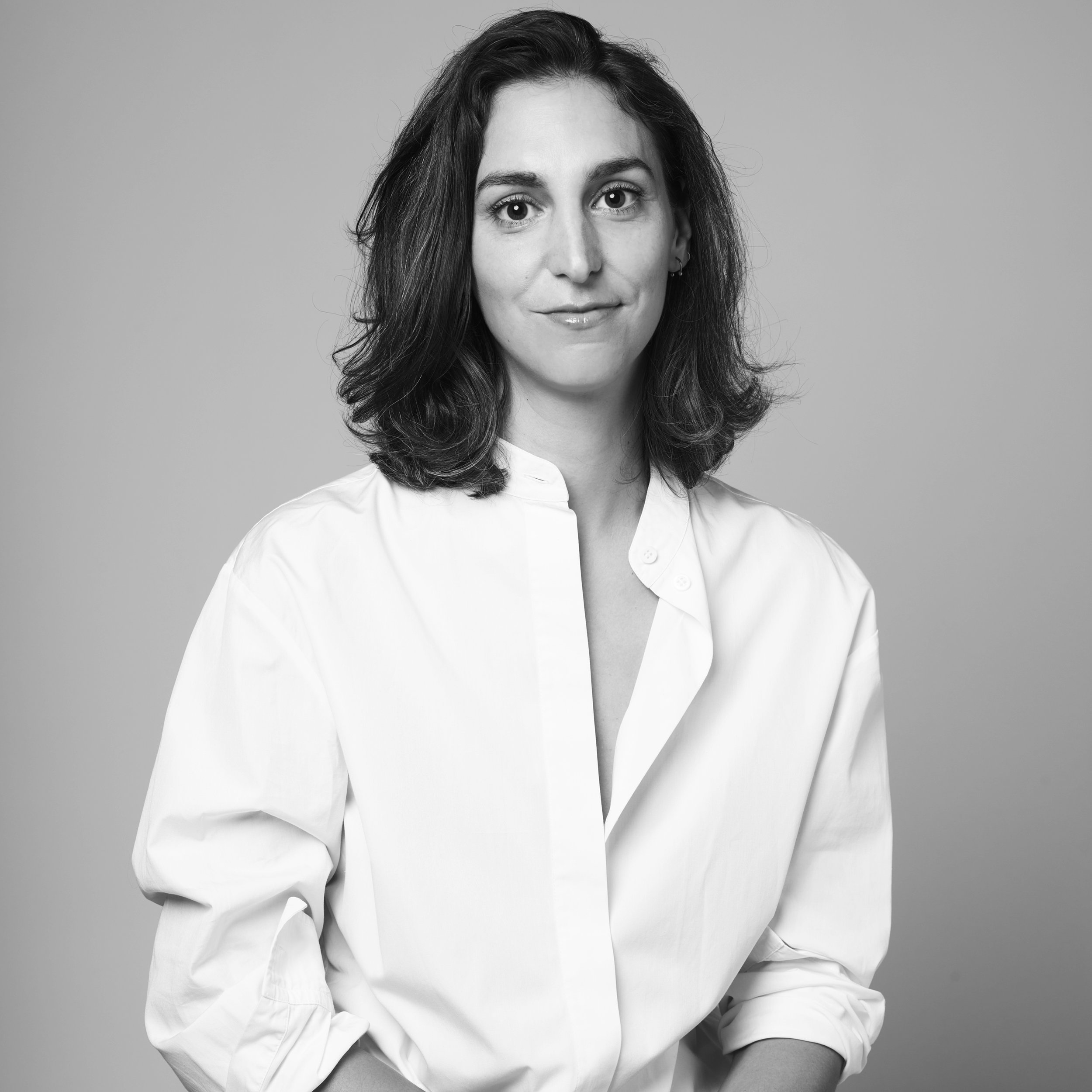 Black and white portrait of a woman with shoulder-length wavy hair, wearing a white shirt with open collar, looking directly at the camera with a slight smile, against a plain light gray background.