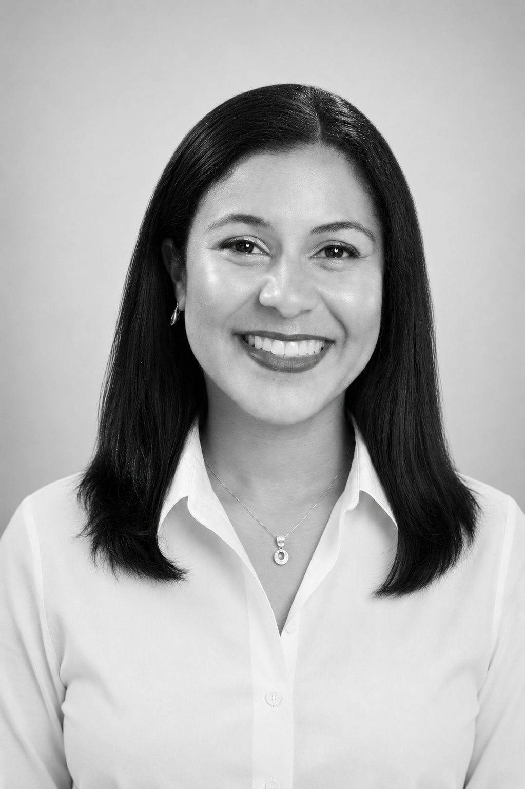 Black and white portrait of a smiling woman with straight, shoulder-length hair, wearing a white collared shirt, earrings, and a necklace, against a plain background.