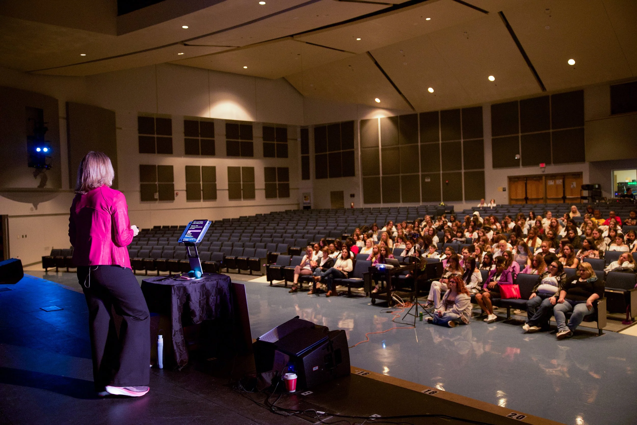 Ellen Claire van Olden on stage speaking to an audience in a large auditorium. The audience includes many women, and the program is Empower the Girls.