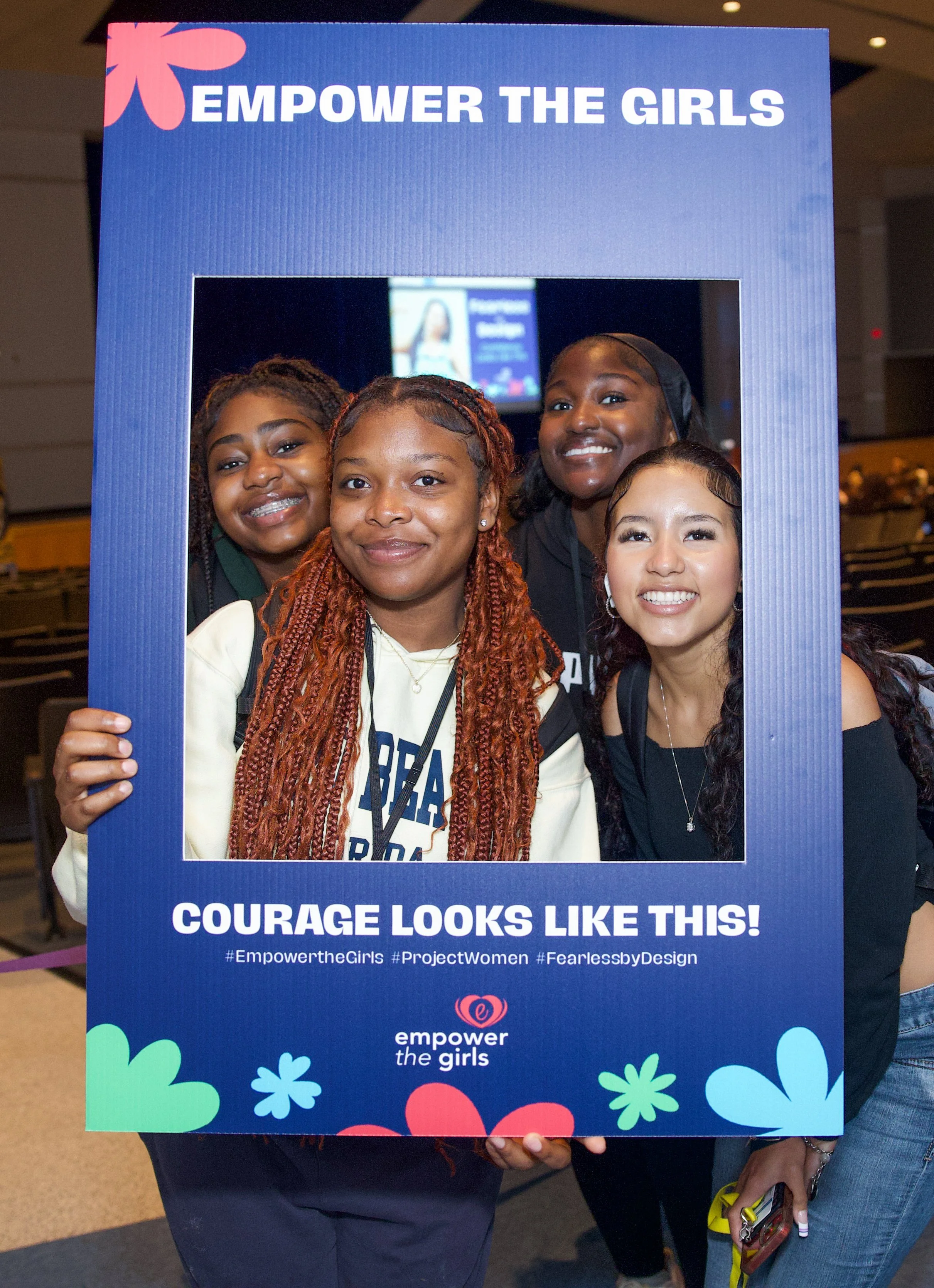 A group of four young women smiling and holding a large blue frame with the text 'Empower the Girls' and the hashtag '#EmpowertheGirls' along with the phrase 'Courage Looks Like This!'.
