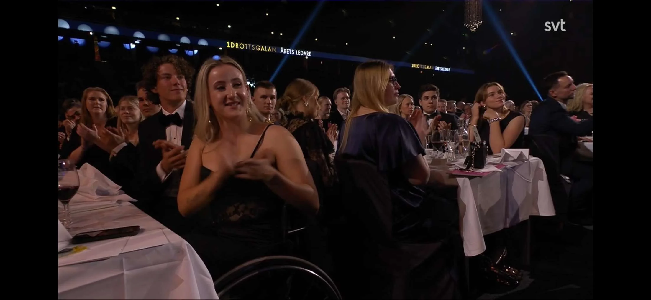 Människor i elegant klädsel sitter vid ett bord på en prisutdelning eller gala. En kvinna i mittbilden klappar, andra personer tittar på scenen.
