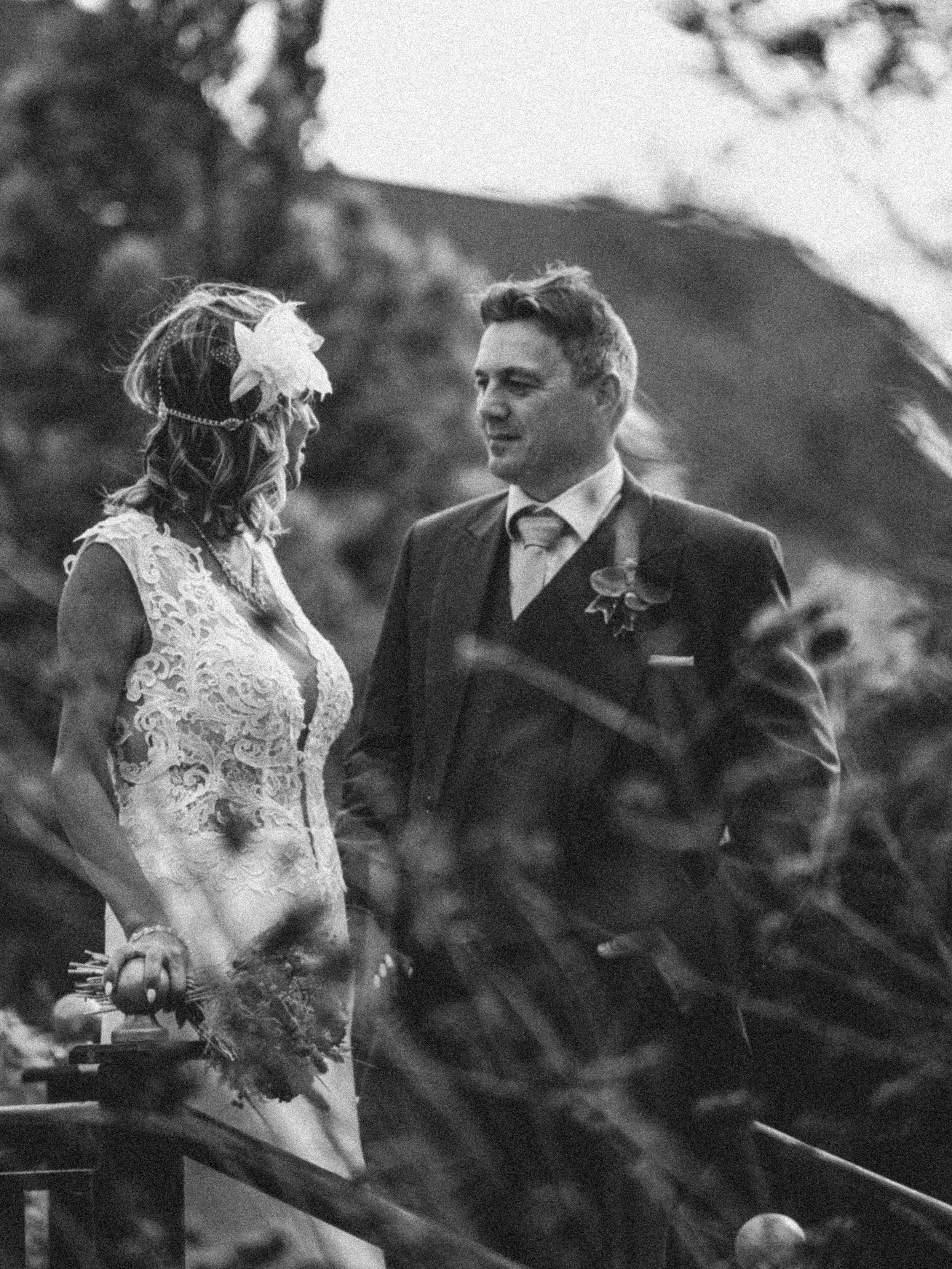 A black and white photograph of a couple dressed in wedding attire, standing on a wooden bridge surrounded by foliage. The woman is wearing a lace wedding dress and a decorative headpiece with feathers, while the man is in a suit with a boutonniere. 