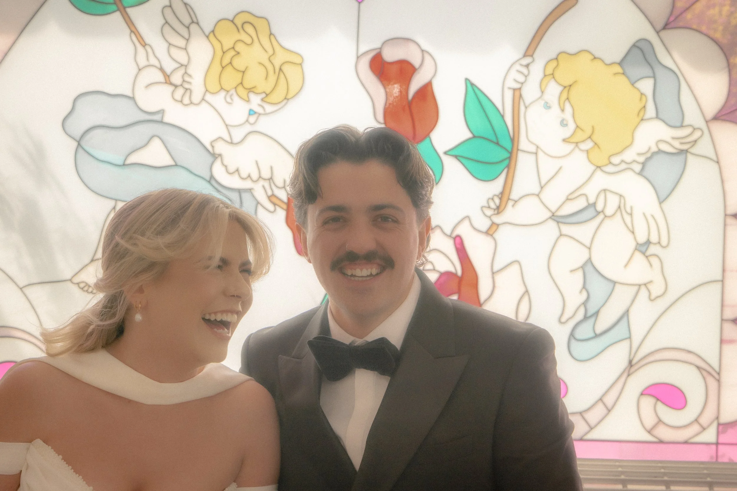 A smiling woman in a white dress with a pearl earring and a man in a tuxedo with a bow tie, both at a formal event, with a colorful illustration of cherubs in the background.