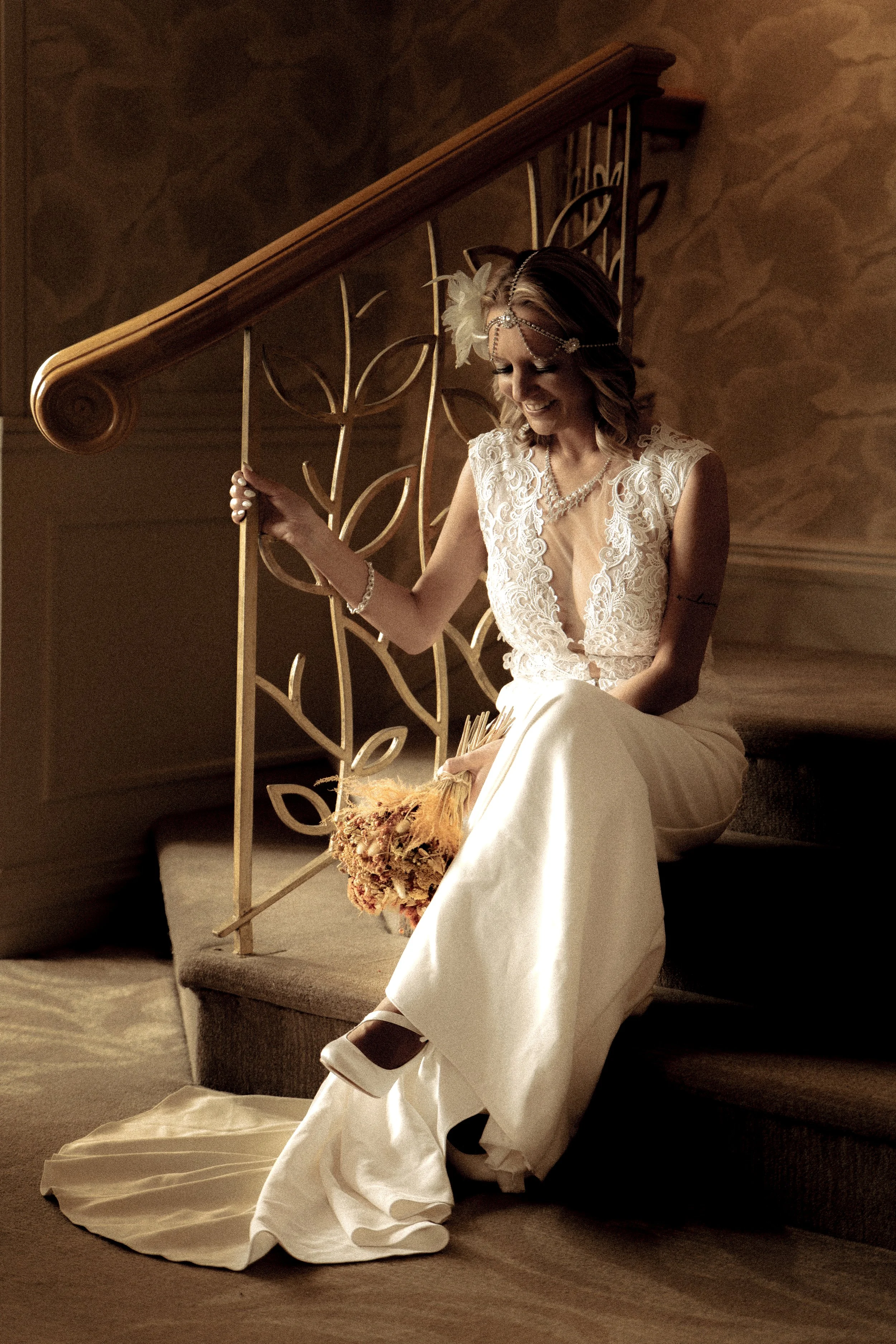 A woman in a white lace dress and veil sitting on stairs, holding a bouquet and smiling.