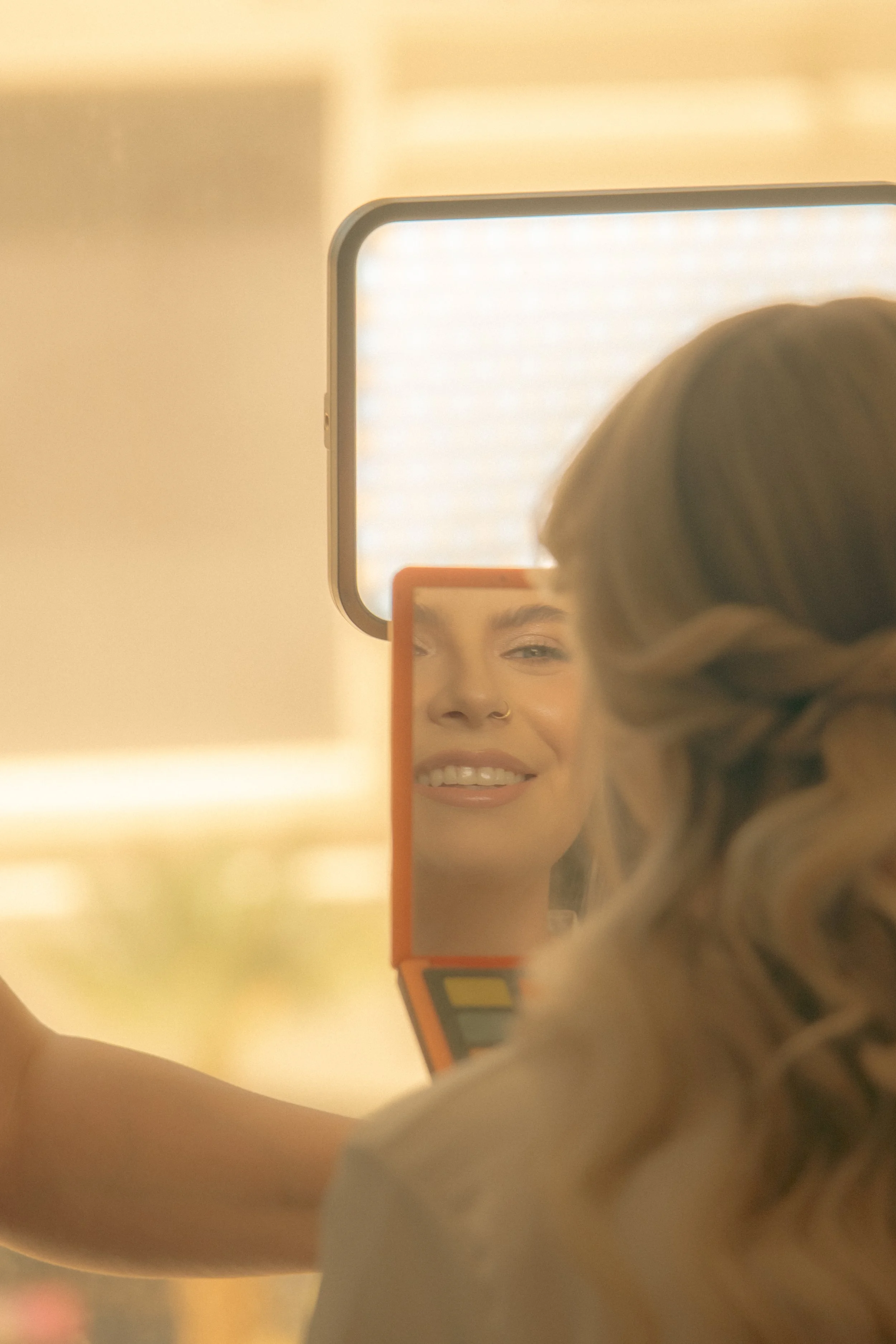 A person looking into a mirror, taking a selfie with a smartphone. The person has brown hair and a nose ring, and is smiling.