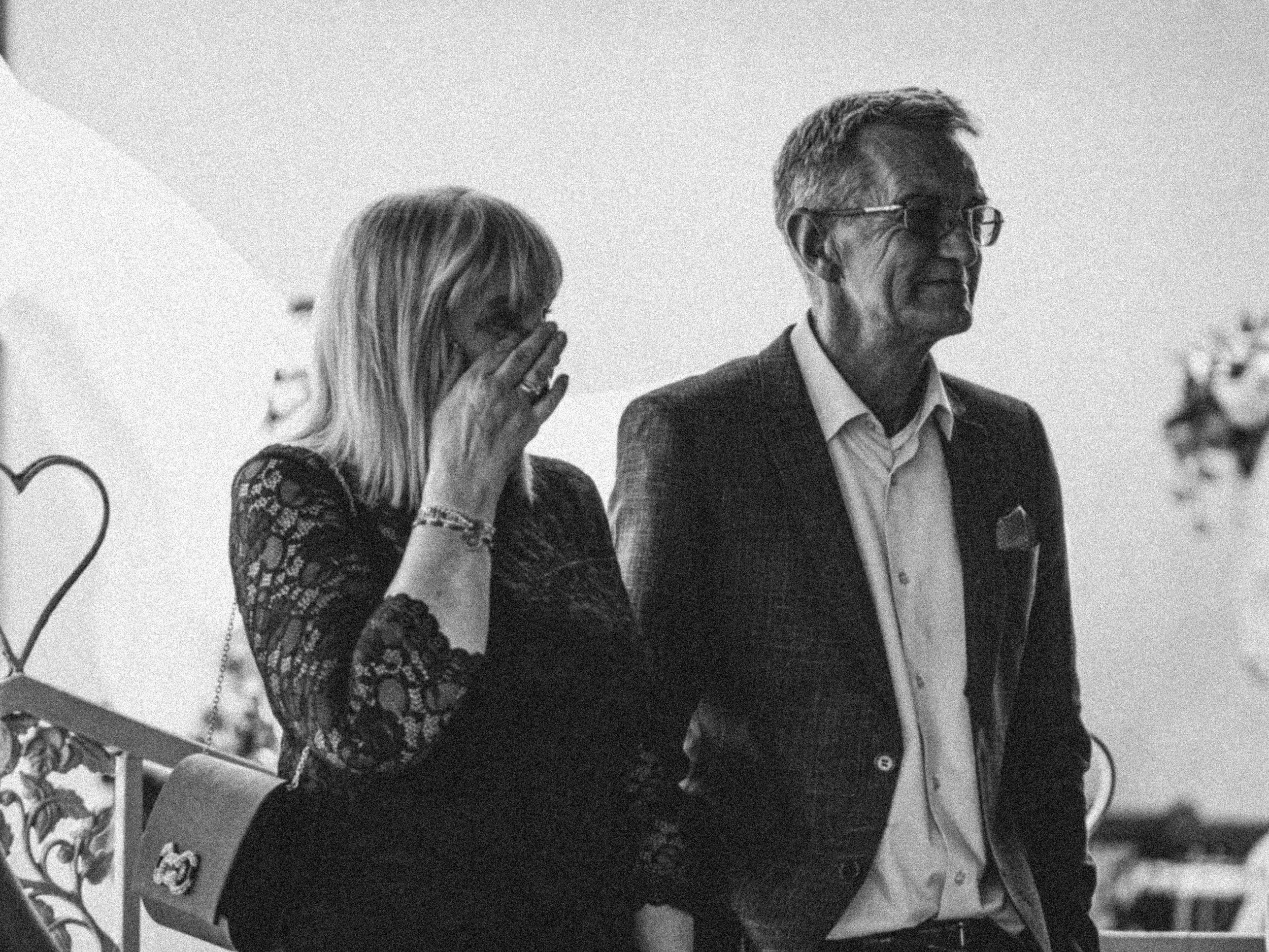 A woman with long hair and a lace top is sitting on a bench, covering her face with her hand, and an older man with glasses is standing beside her, dressed in a suit and smiling.