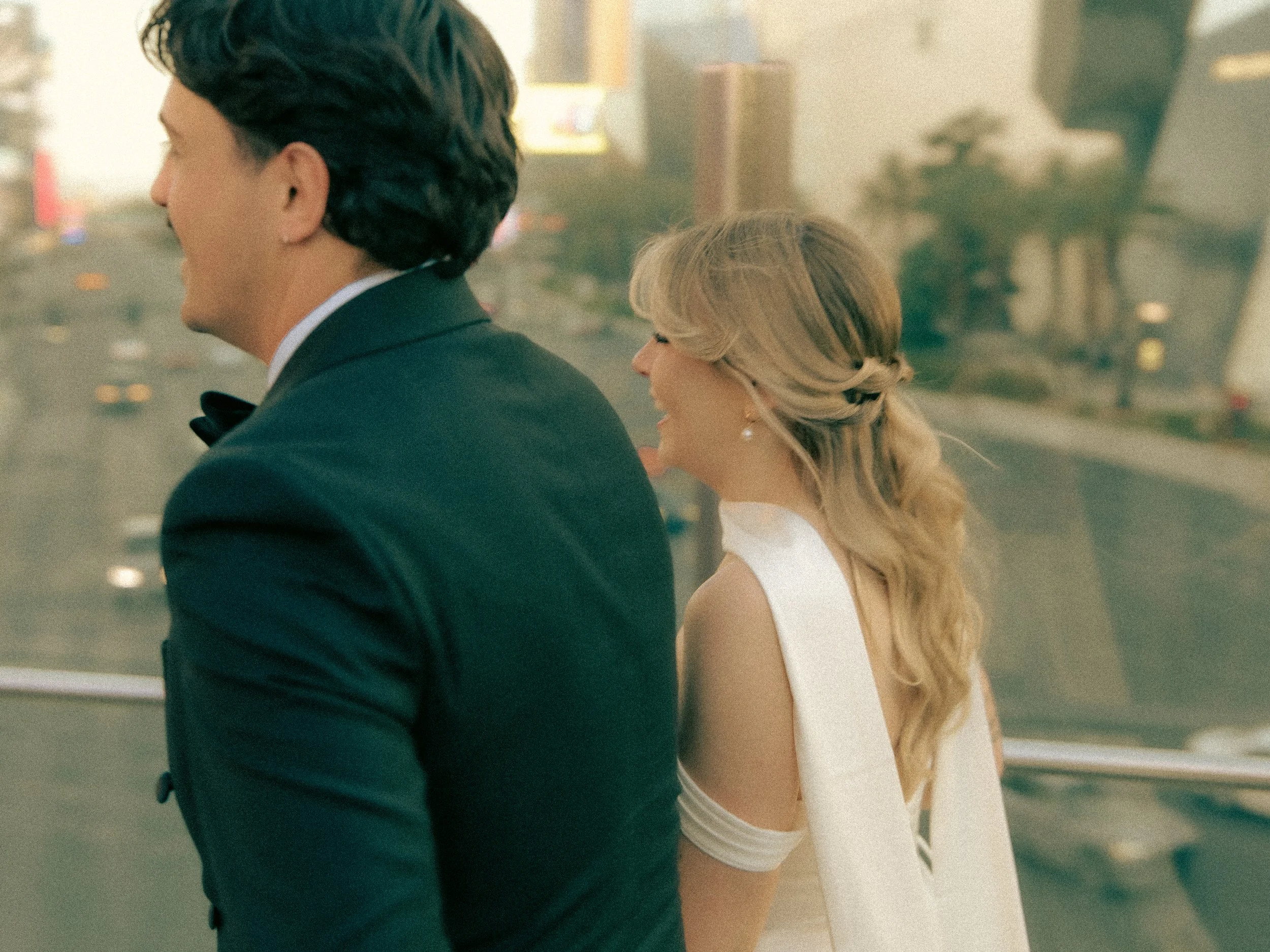 A man and woman dressed formally, standing close together, with city buildings and traffic blurred in the background.
