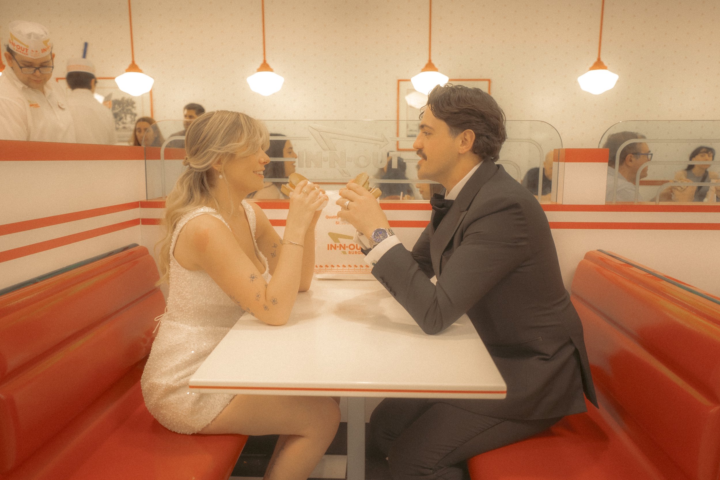 A couple is sitting at a booth in a restaurant, facing each other and holding small sandwiches, smiling and looking into each other's eyes. The woman has long blonde hair and tattoos on her arm, wearing a white dress, and the man has dark curly hair,