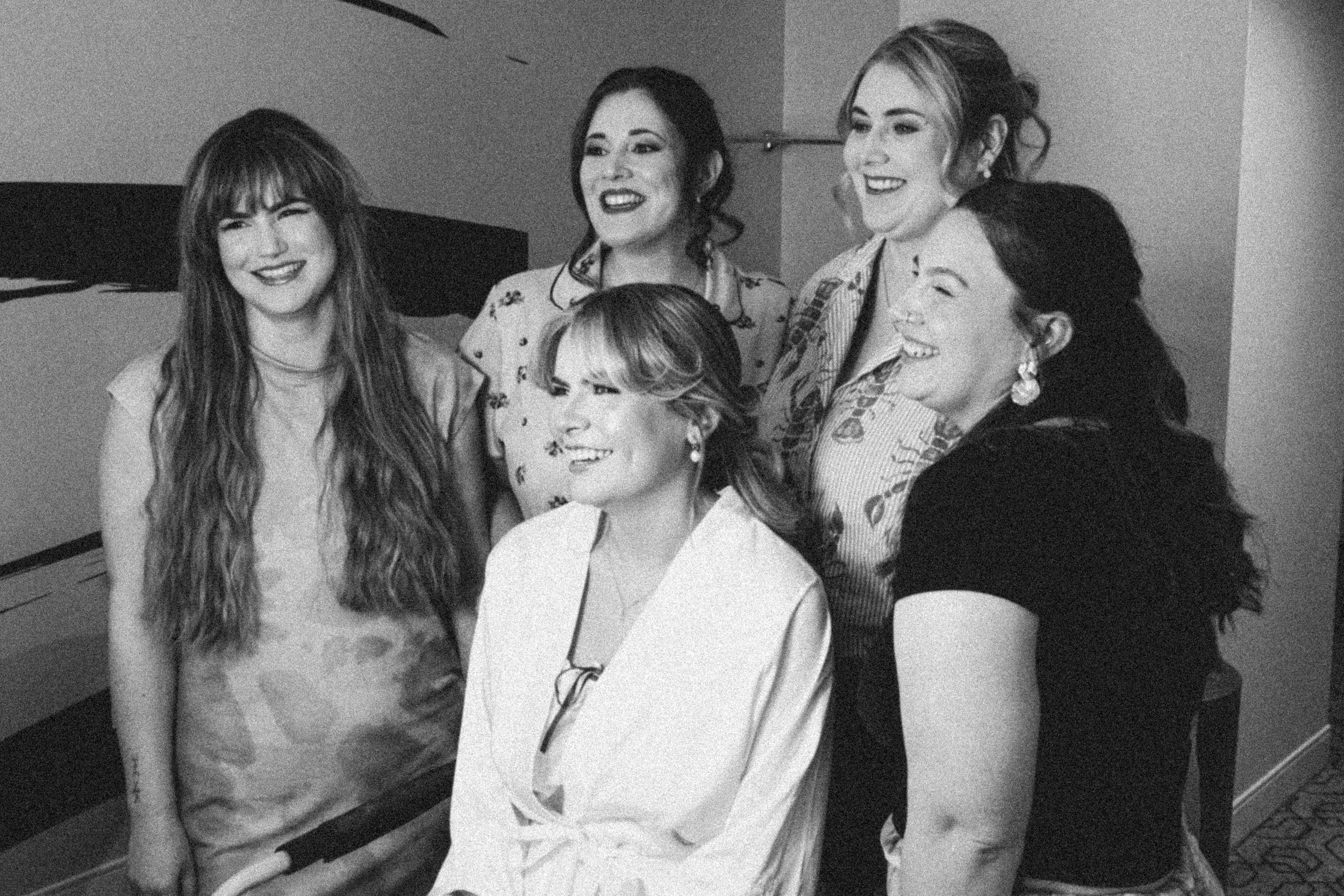 Five women are smiling and looking at the camera in an indoor setting, with some standing and some seated.