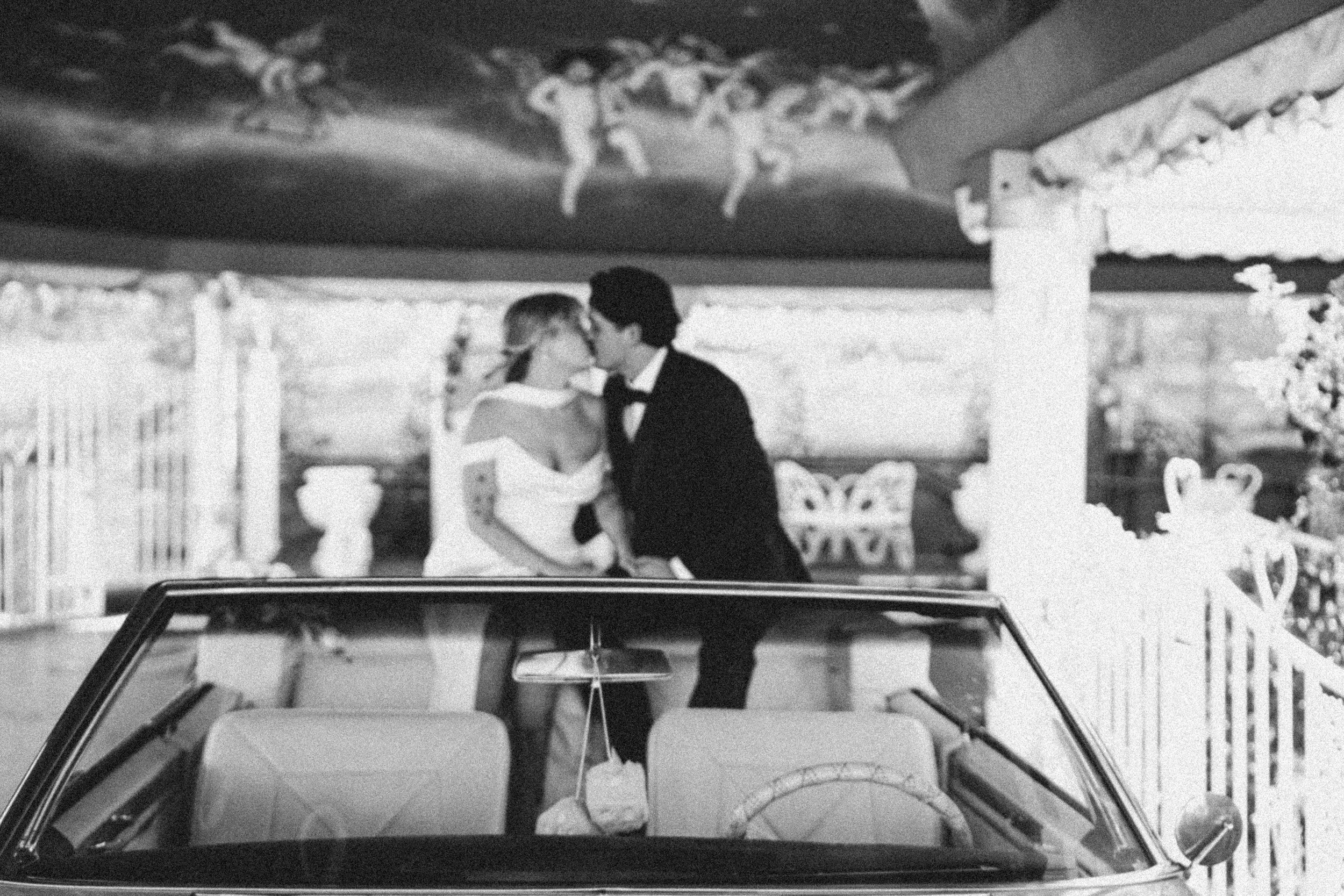 A black and white photo of a couple kissing in front of a vintage convertible car. The couple is dressed formally, and the background shows a decorative area with a white fence and flowers. The scene appears romantic and celebratory.